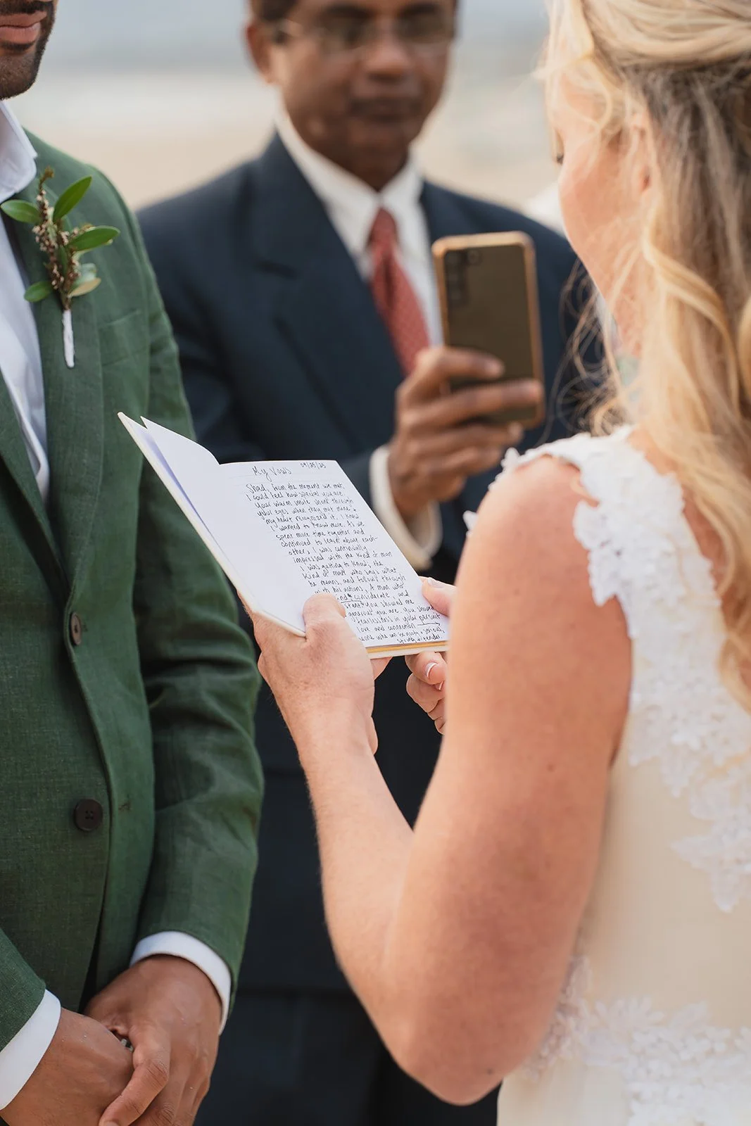 A bride and groom exchange vows during their wedding ceremony, with the bride reading from a small book and the groom listening, while an officiant records the moment with a smartphone.