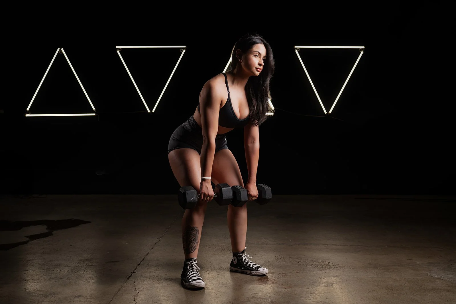 A woman in black workout clothes doing a bent-over dumbbell exercise in a dark gym with illuminated triangular light fixtures in the background.