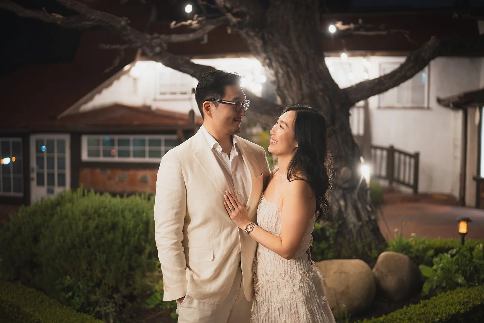 A couple dressed elegantly in formal attire standing close together outdoors at night, gazing into each other's eyes with smiles, with a large tree and warm lights in the background.
