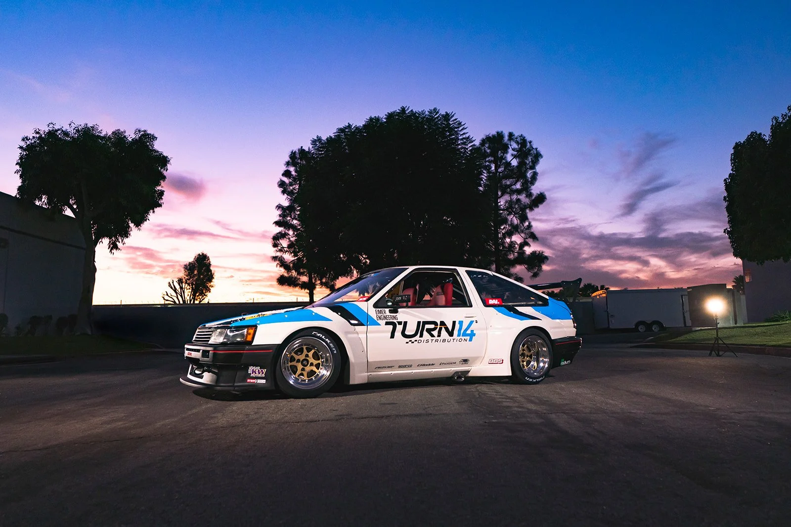 A vintage race car with a white, blue, and black wrap featuring sponsors, parked on a paved surface at sunset, with trees and trailers in the background.