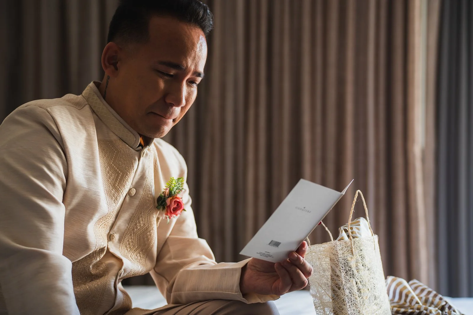 A groom in traditional attire sitting on a bed, reading a card with wedding gifts and a decorative bag nearby.