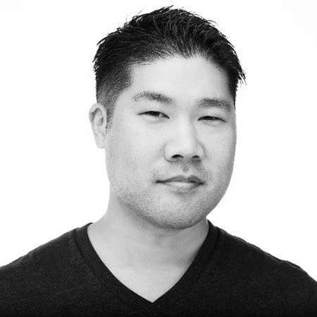 Black and white portrait of a young Asian man with short, dark hair, wearing a black V-neck shirt, looking at the camera with a neutral expression.