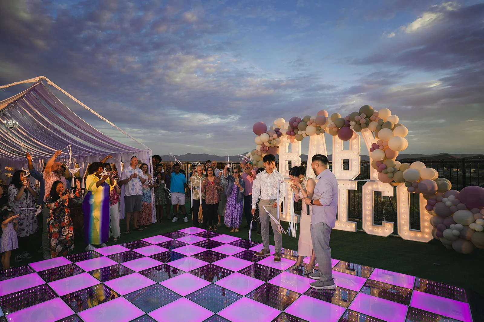 Celebration at sunset with a group of people dancing on a glowing pink and purple checkered dance floor, large illuminated letters 'H' and 'Q' decorated with balloons, and a purple tent in the background.