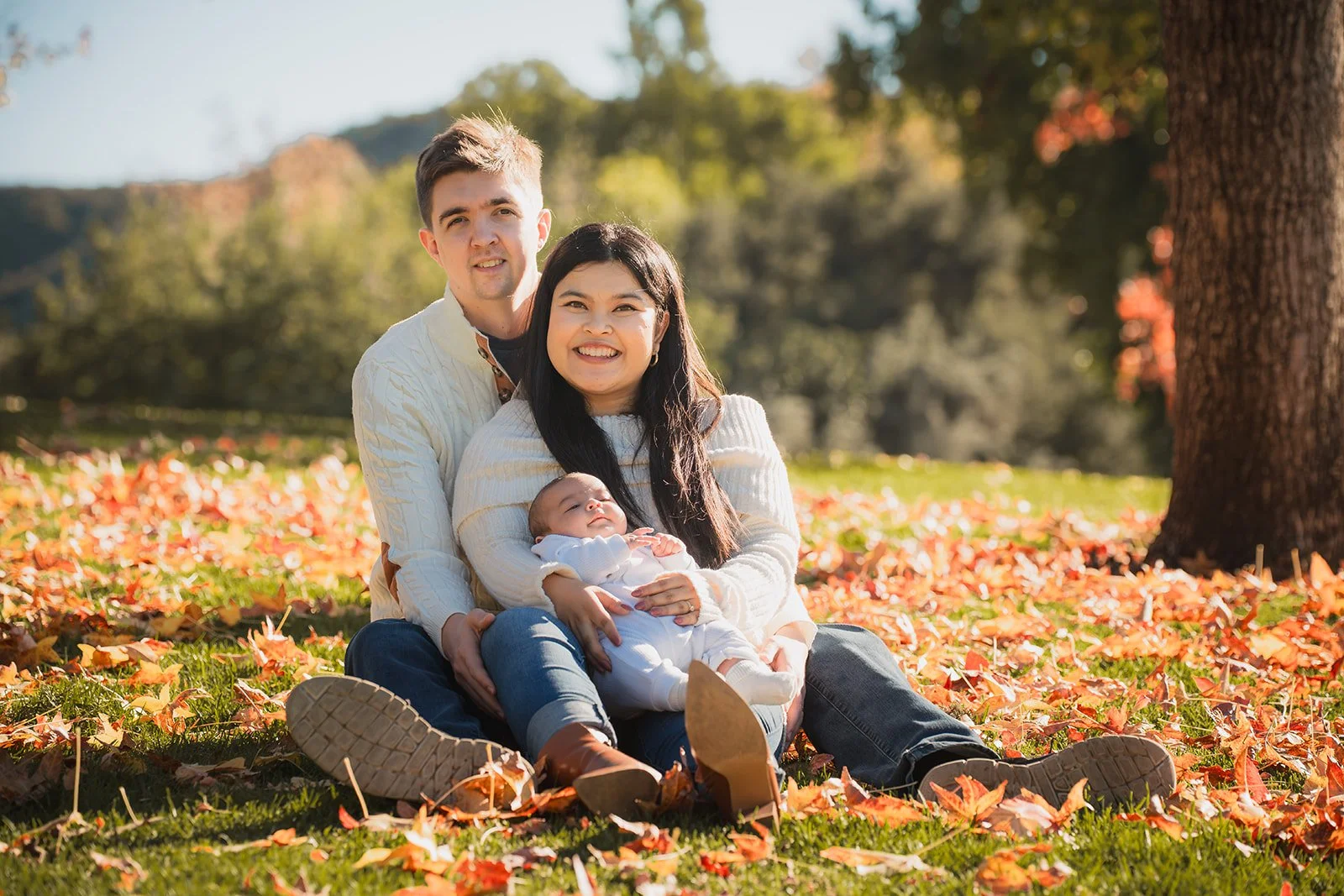 A happy family of three, a man, a woman, and a baby, sitting on the grass amidst fallen autumn leaves outdoors, smiling at the camera, with trees and a clear sky in the background.