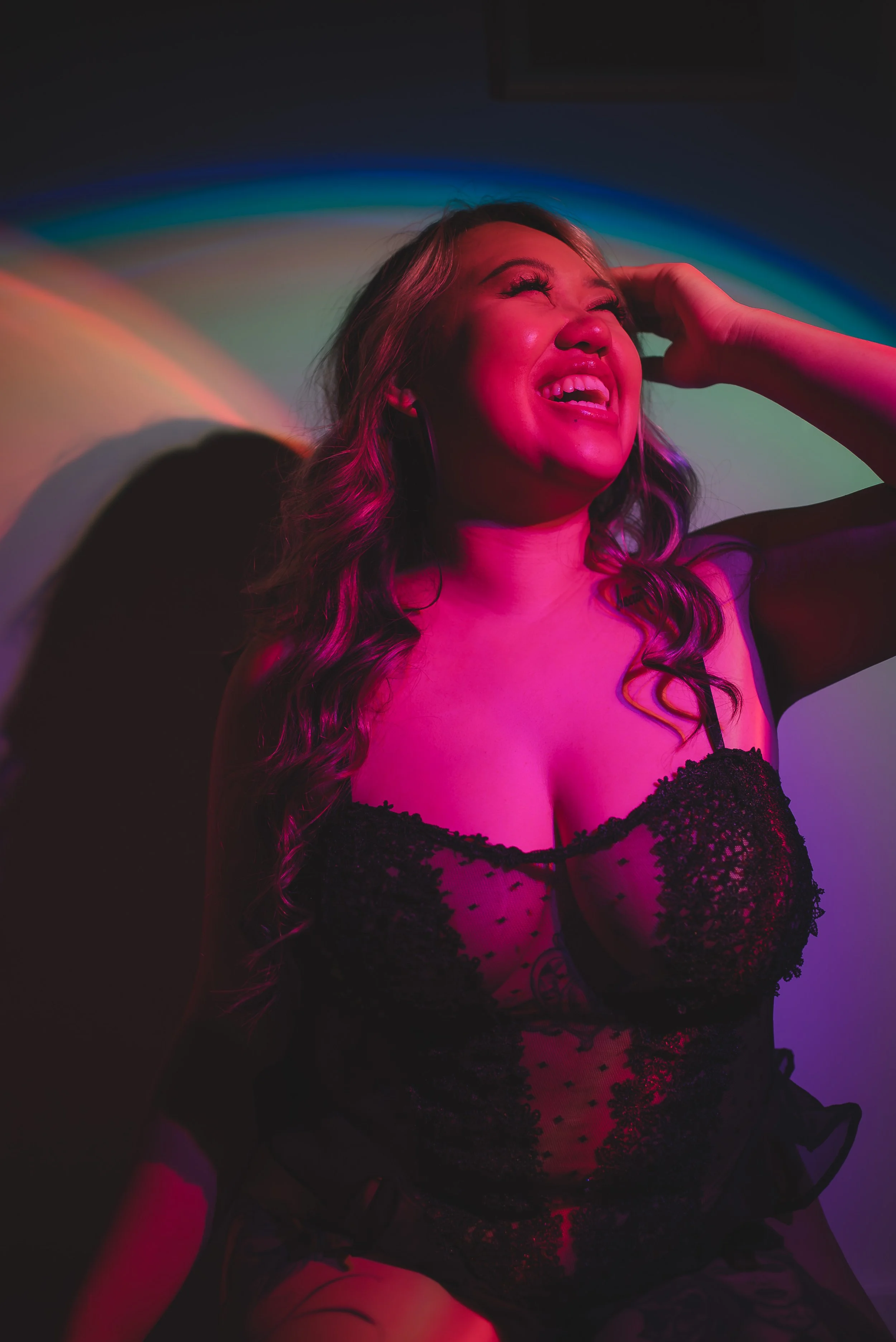 A woman with long wavy hair, wearing a black lace dress, is smiling and laughing with her head tilted back, illuminated by pink and colorful lights.