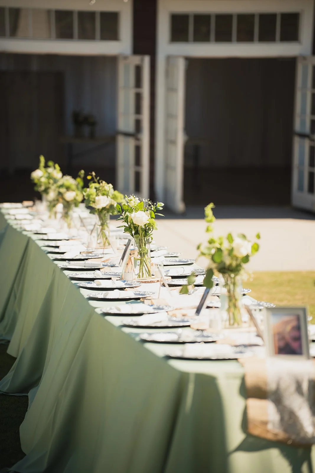 Long banquet table decorated with vases of green and white flowers, set with plates and glasses, outdoors in front of a barn or rustic building.