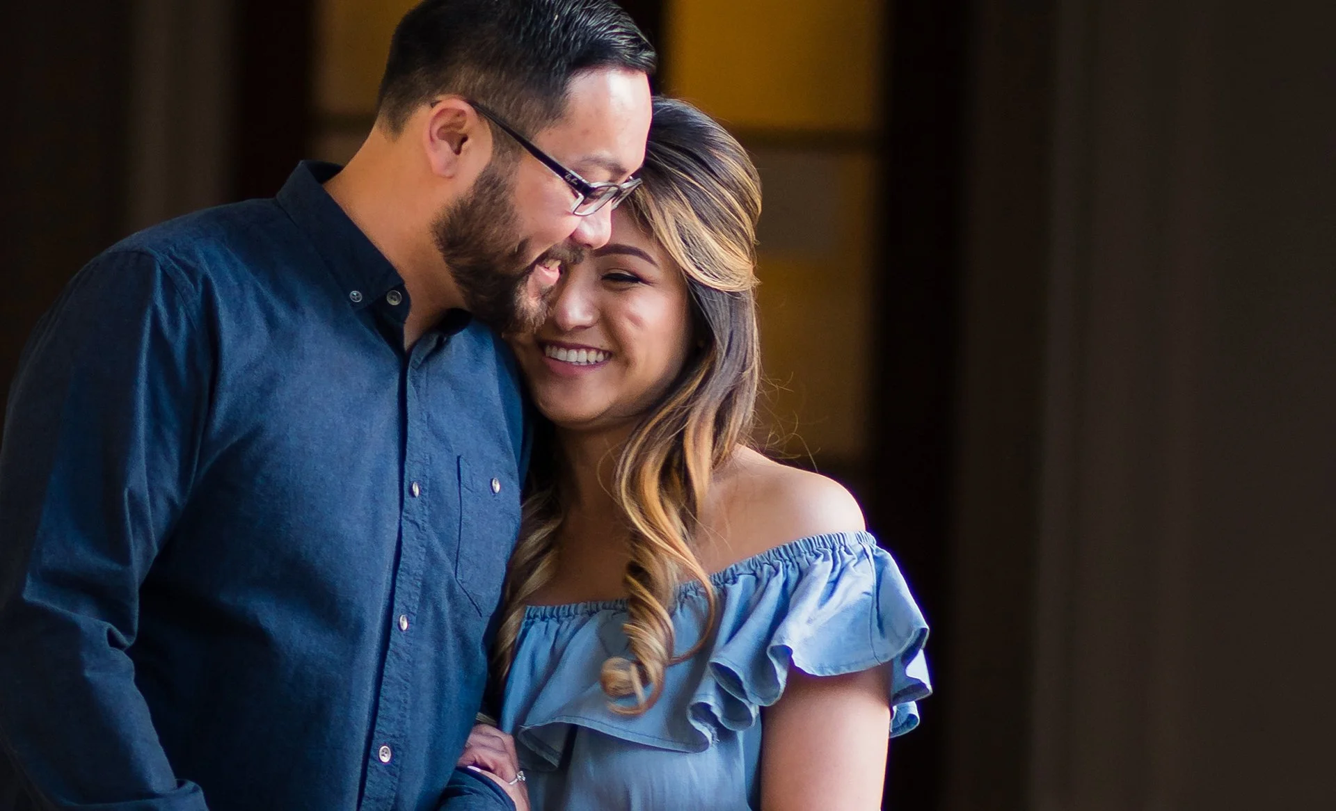 A happy couple sharing an intimate moment, smiling and leaning their foreheads together, with warm indoor lighting in the background.
