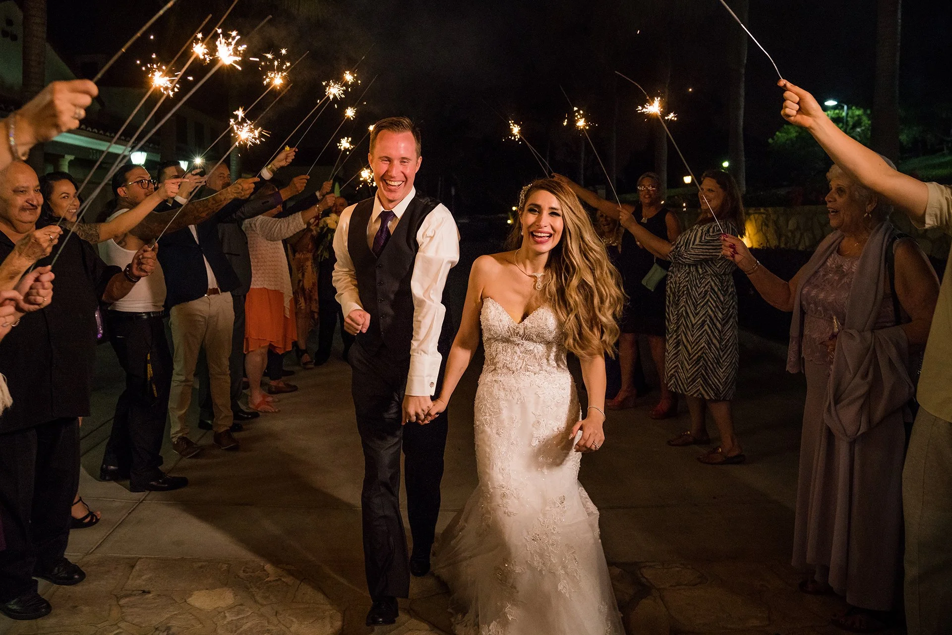 A newlywed couple walks hand-in-hand through a nighttime reception, surrounded by guests holding sparklers, celebrating their wedding.