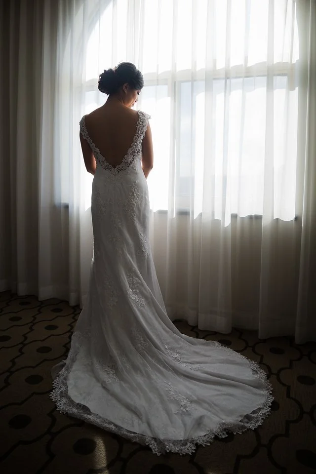 A bride in a white lace wedding gown standing in front of a window with sheer curtains, looking away from the camera.