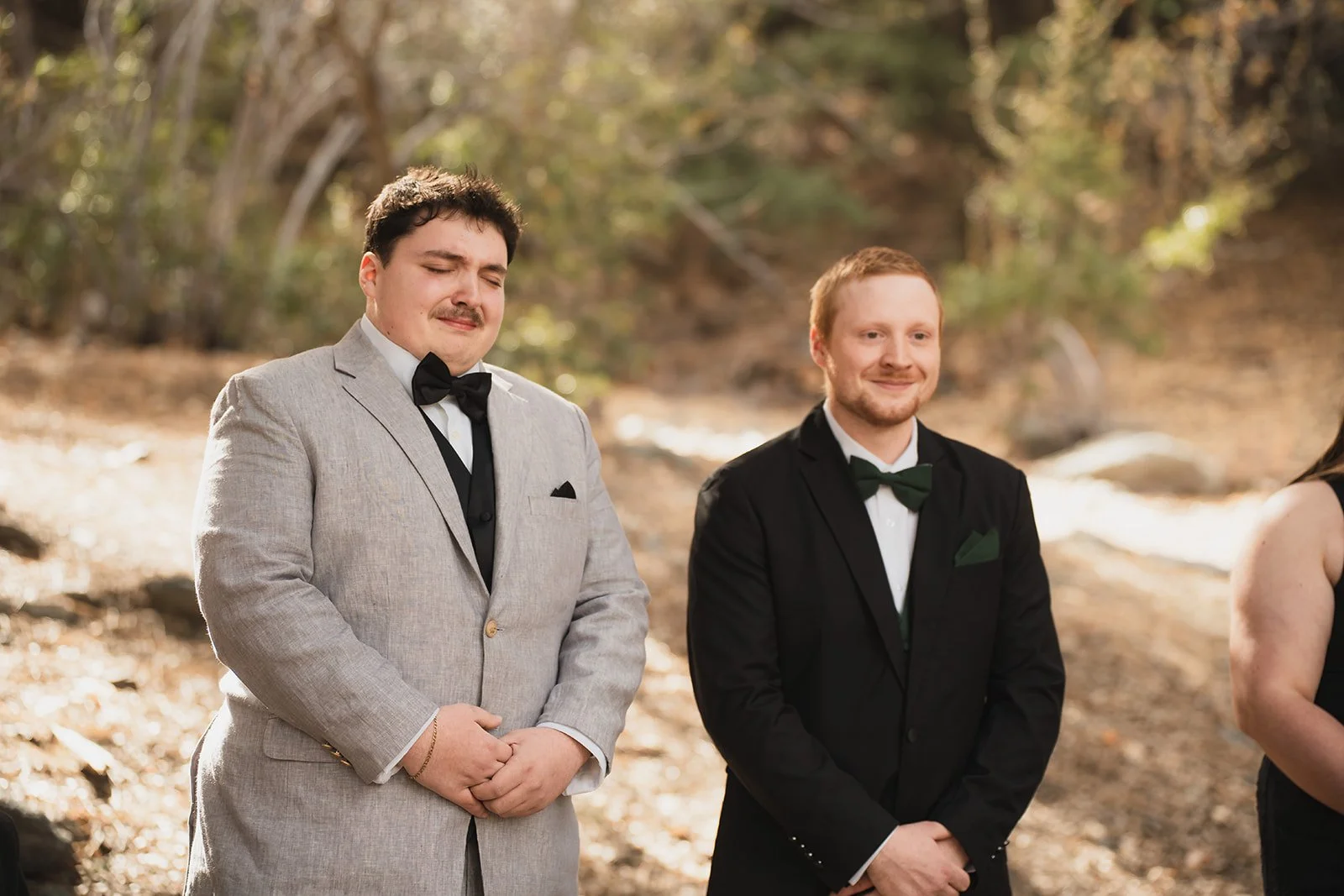 Two men in tuxedos standing outdoors in a wooded area, with expressions of solemnity or prayer, participating in a ceremony or event.