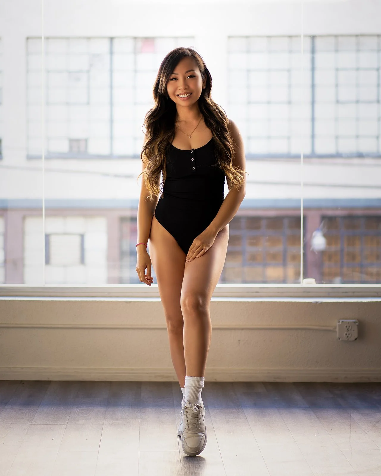 Young woman with long, wavy hair wearing a black tank top, white sneakers, and socks, standing inside in front of a large window with a cityscape view, smiling confidently.