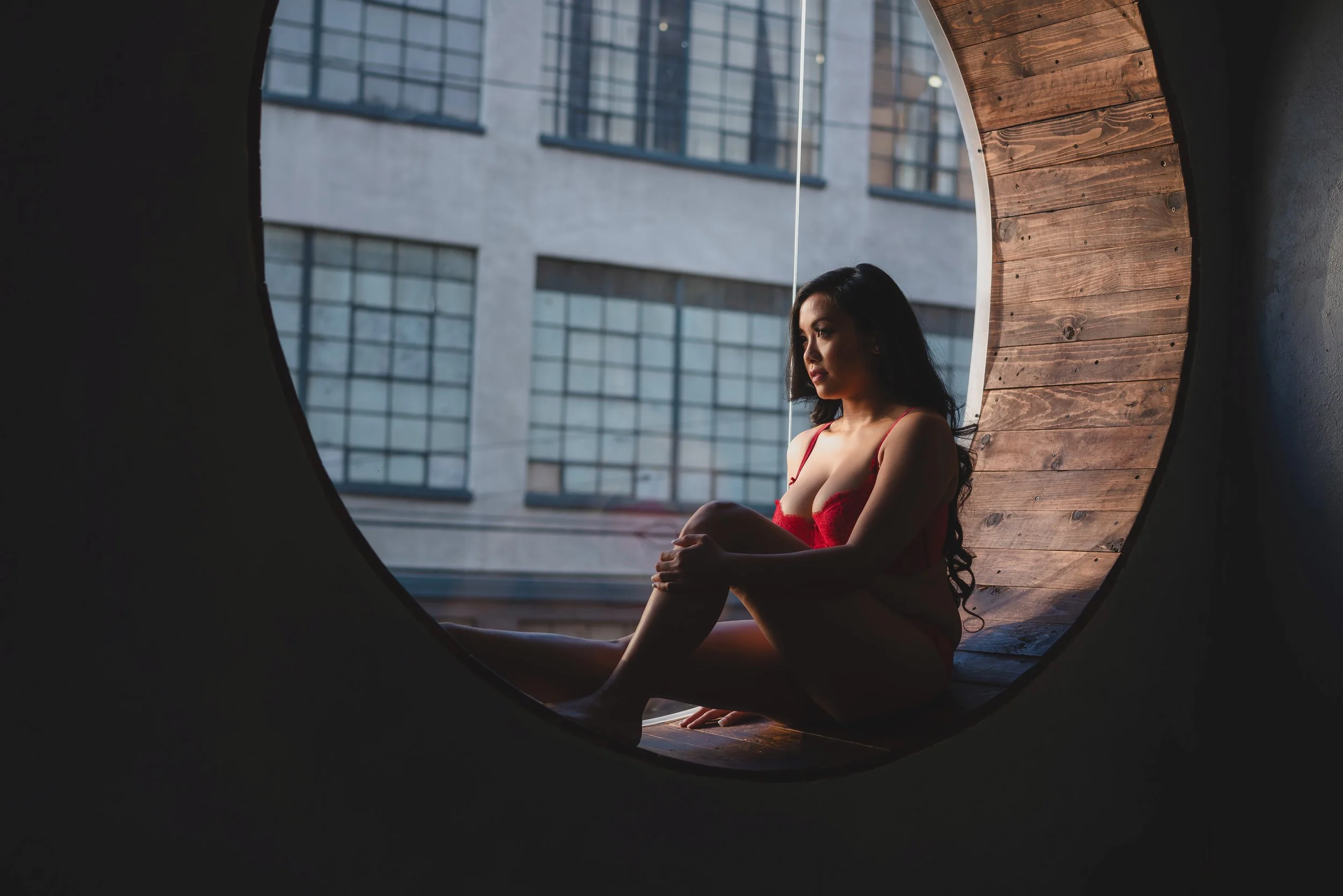 Woman in red lingerie sitting by a circular window with a cityscape view in the background.