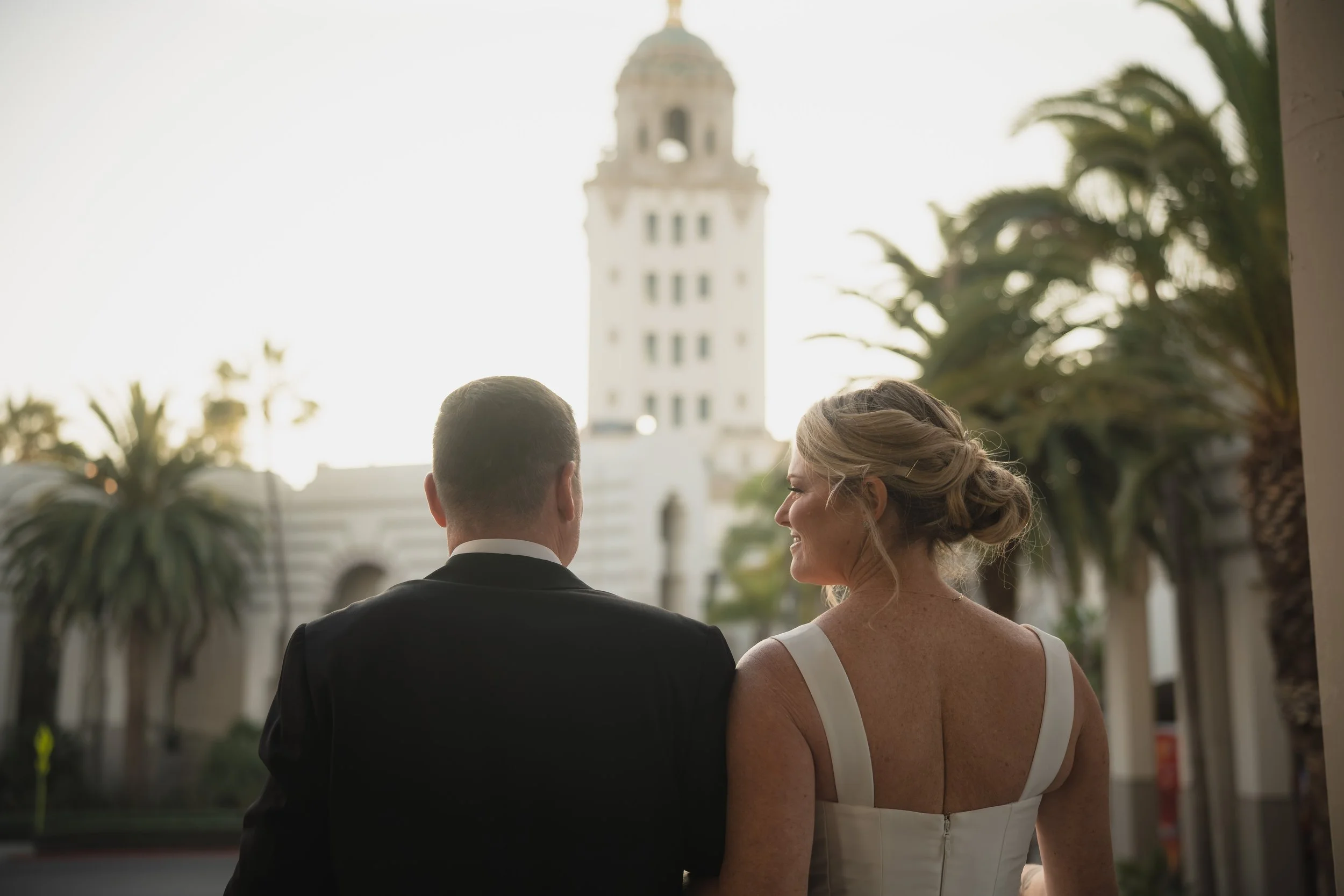 A bride and groom standing together outdoors near a white building and palm trees, facing away from the camera during sunset.