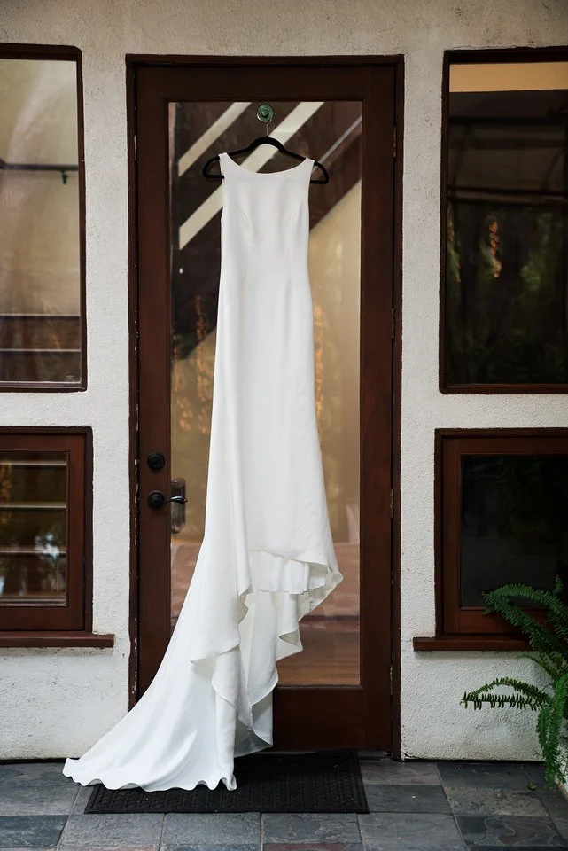A white wedding dress hanging on a black hanger on a door with glass panels, outside on a porch with stone flooring and a potted fern to the side.