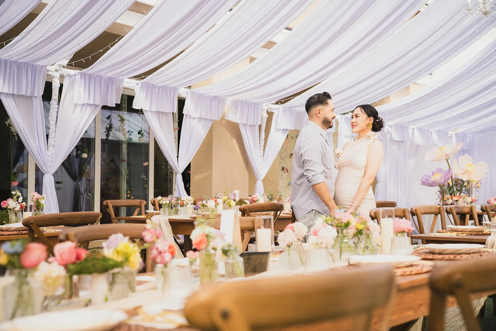 A couple stands face to face under a decorated canopy at a wedding reception, with flower arrangements and table settings visible.