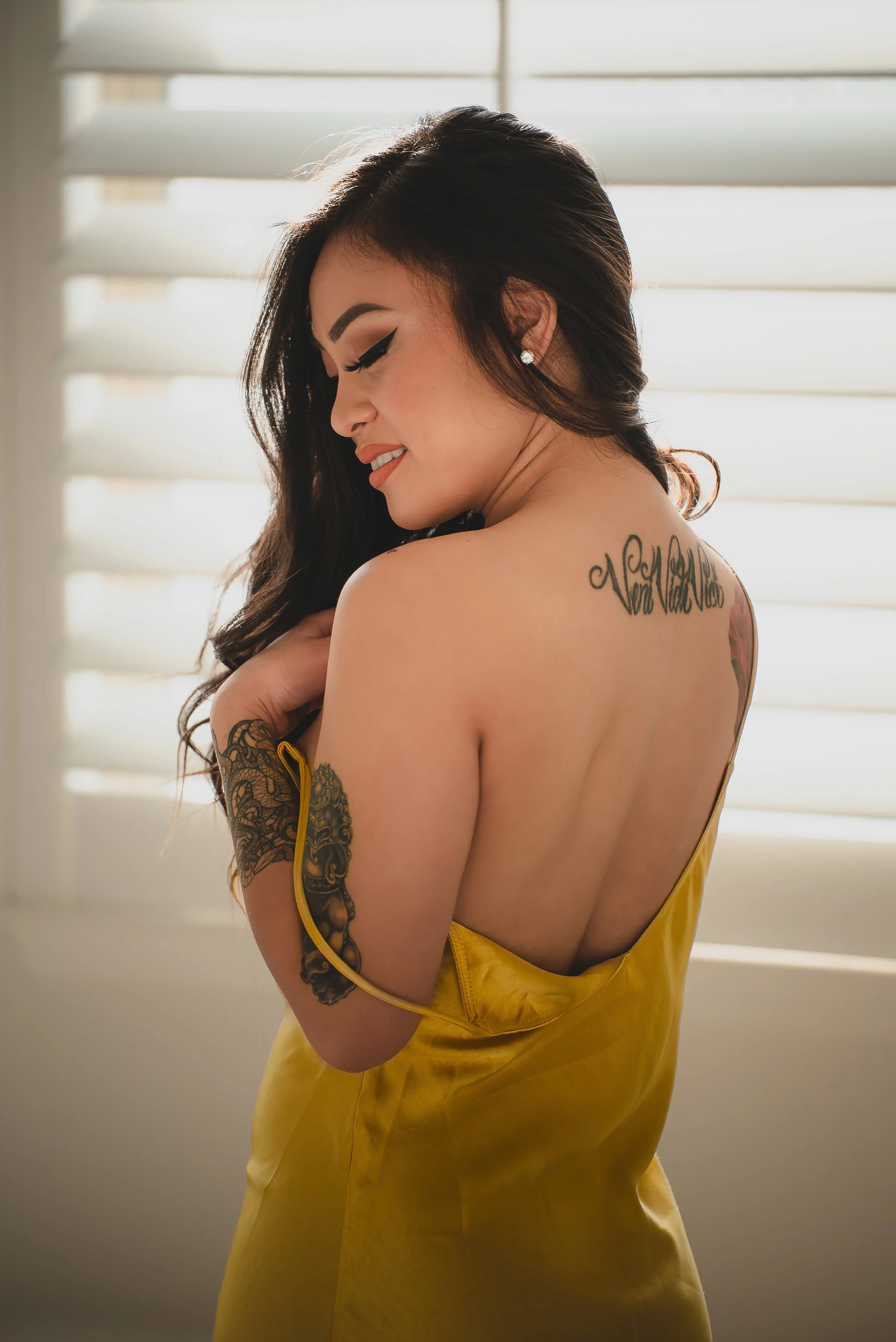 A woman with long dark hair, tattoos on her arm and back, wearing a yellow dress, stands near window blinds, smiling with eyes closed.