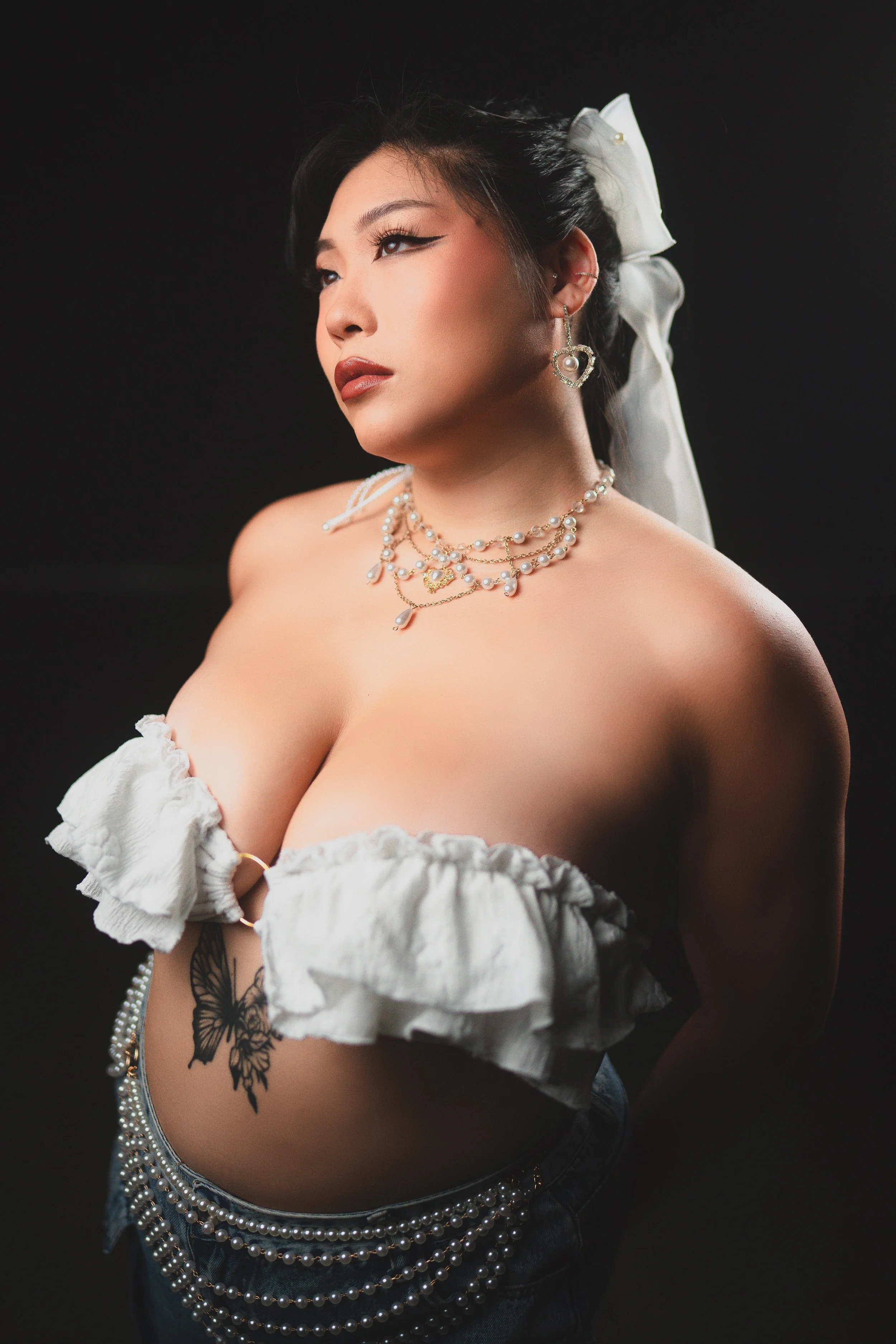 A woman with dark hair in a ponytail, wearing pearl jewelry and a white off-shoulder top, posing against a black background.