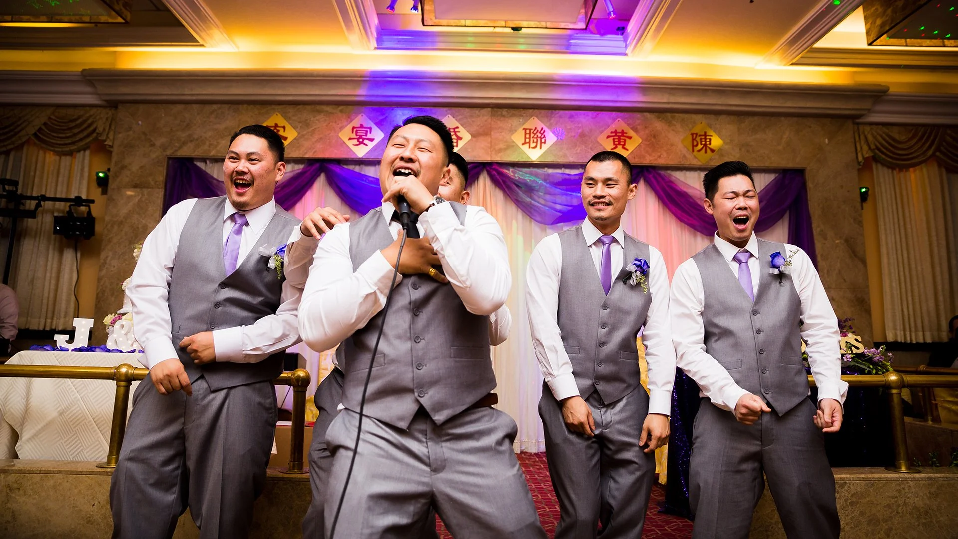 Group of men dressed in formal gray vests and pants, singing and dancing on a decorated stage at a celebration or wedding.