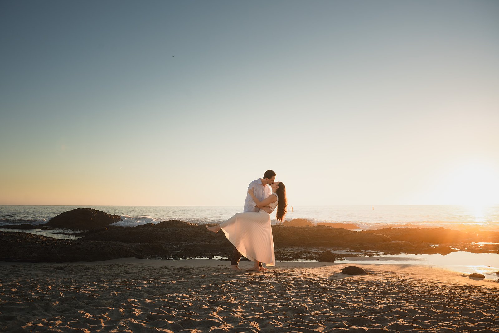 A couple dressed in white sharing a kiss on the beach at sunset, with rocks and water in the background.
