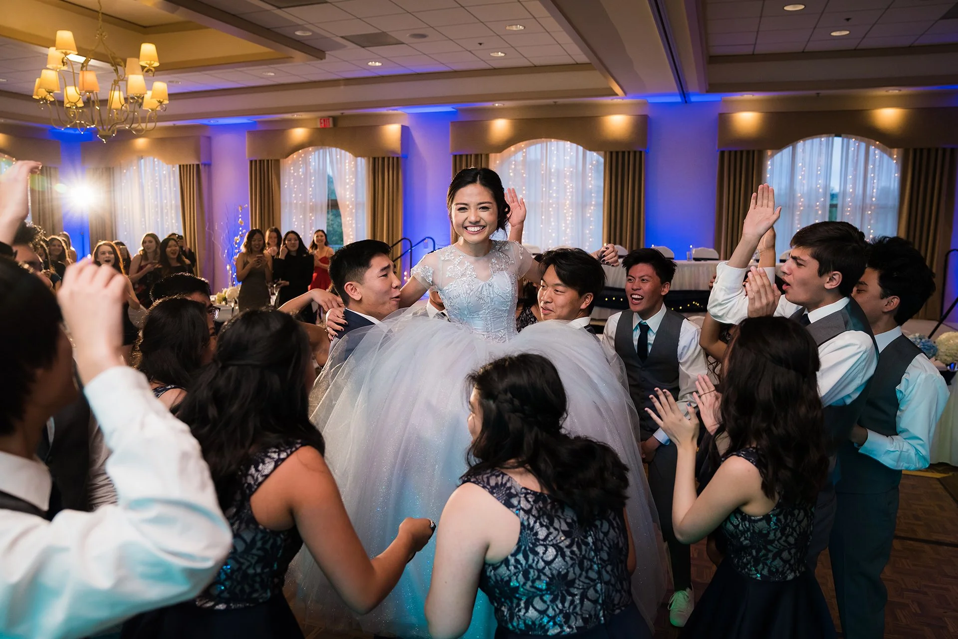 Bride in a white wedding gown being lifted by friends in a decorated banquet hall.