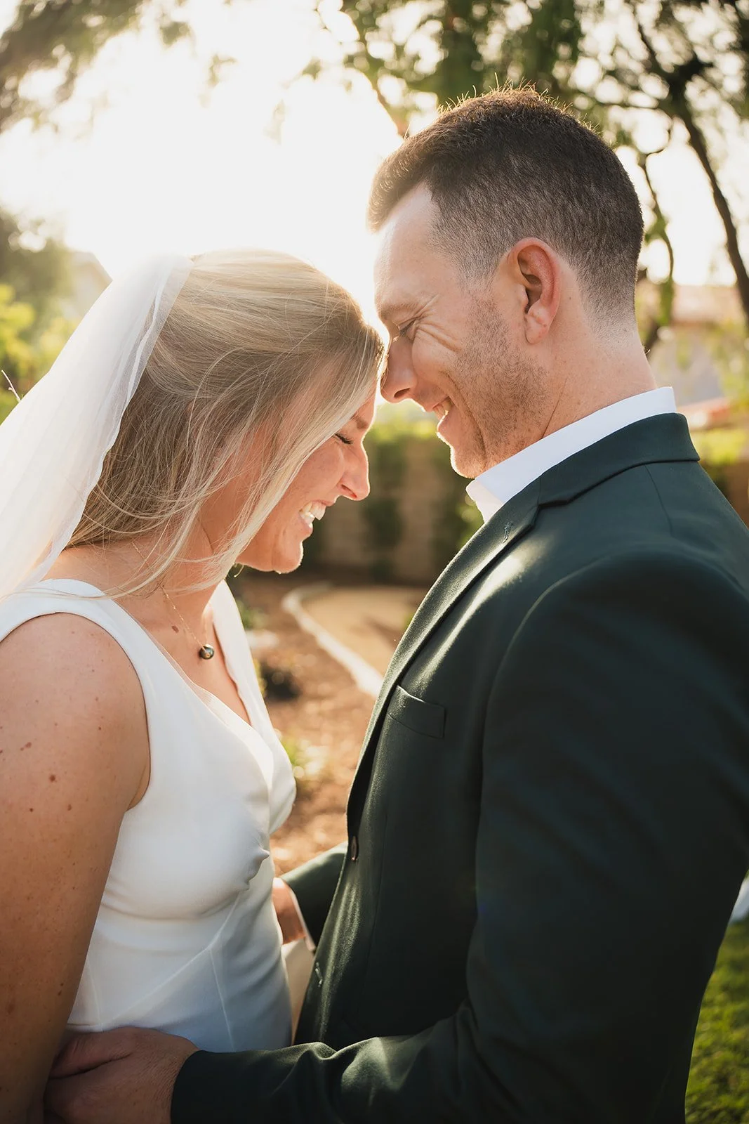 A bride and groom smiling with their foreheads touching outdoors during sunset.