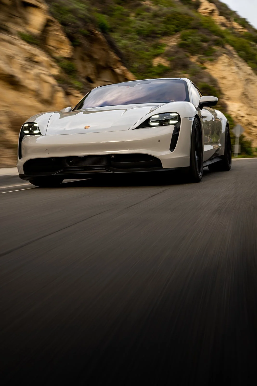 A white sports car, likely a Porsche, driving fast on a winding mountain road with rocky cliffs and green vegetation in the background.