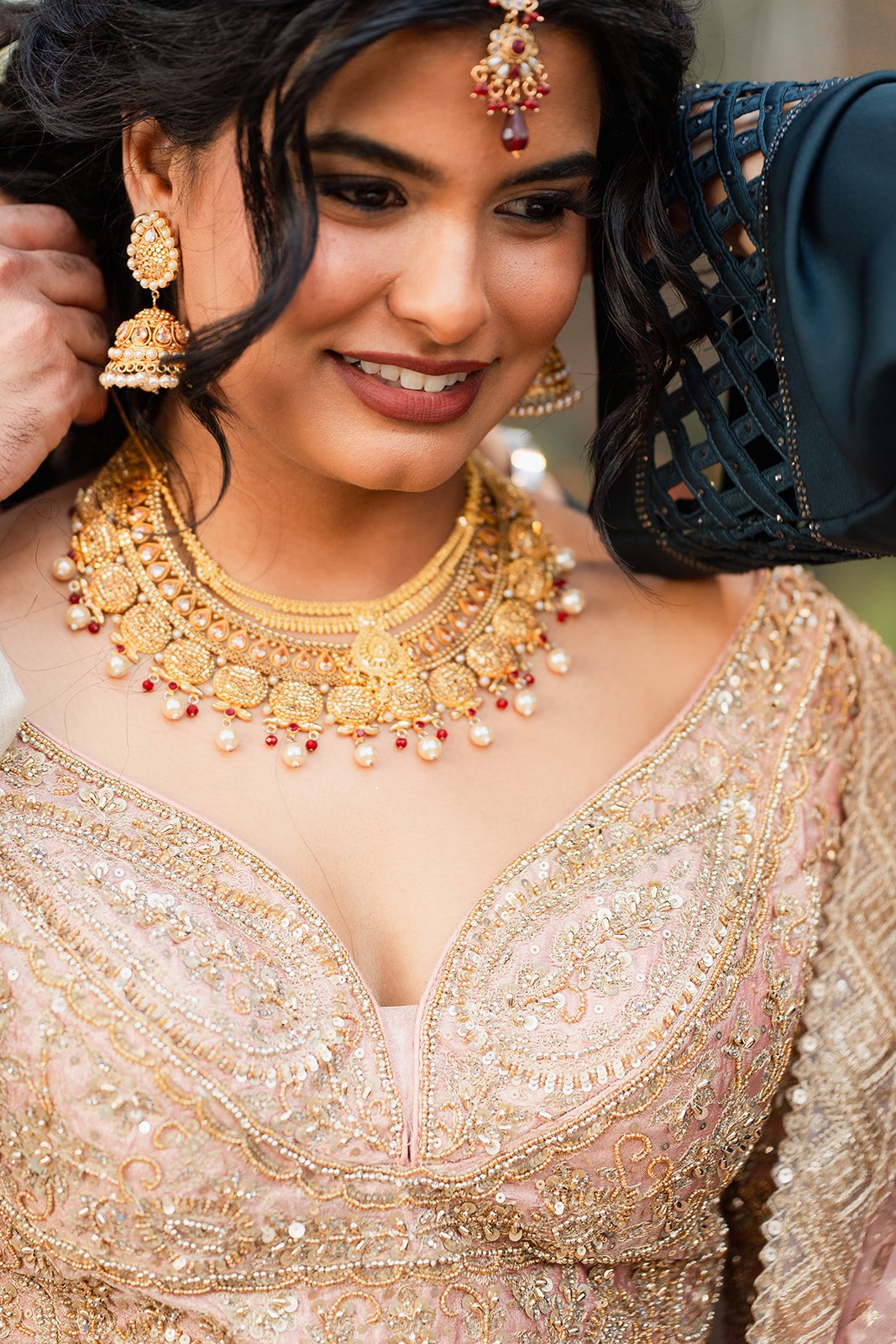A woman dressed in traditional Indian attire wearing elaborate gold and pearl jewelry, including a necklace, earrings, and a maang tikka, smiling gently.