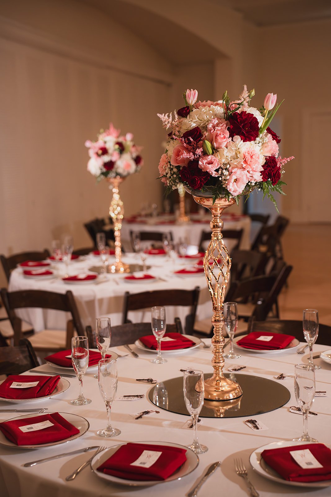 Elegant banquet table setup with white tablecloth, red napkins, and tall floral centerpieces featuring pink, white, and red flowers.