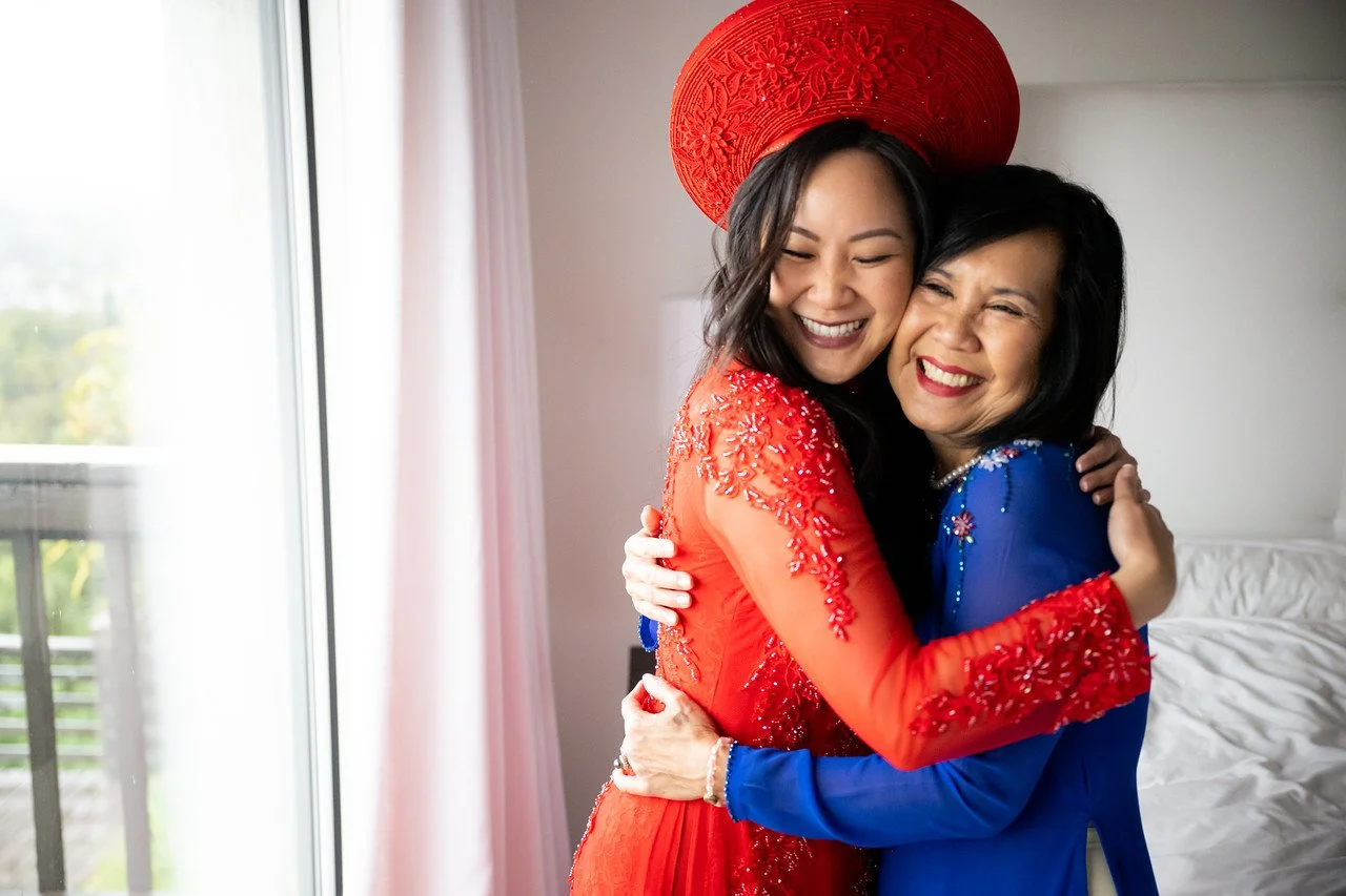Two women hugging each other and smiling widely, one wearing a red dress and large red hat, the other in a blue dress, inside a home near a window with pink curtains.