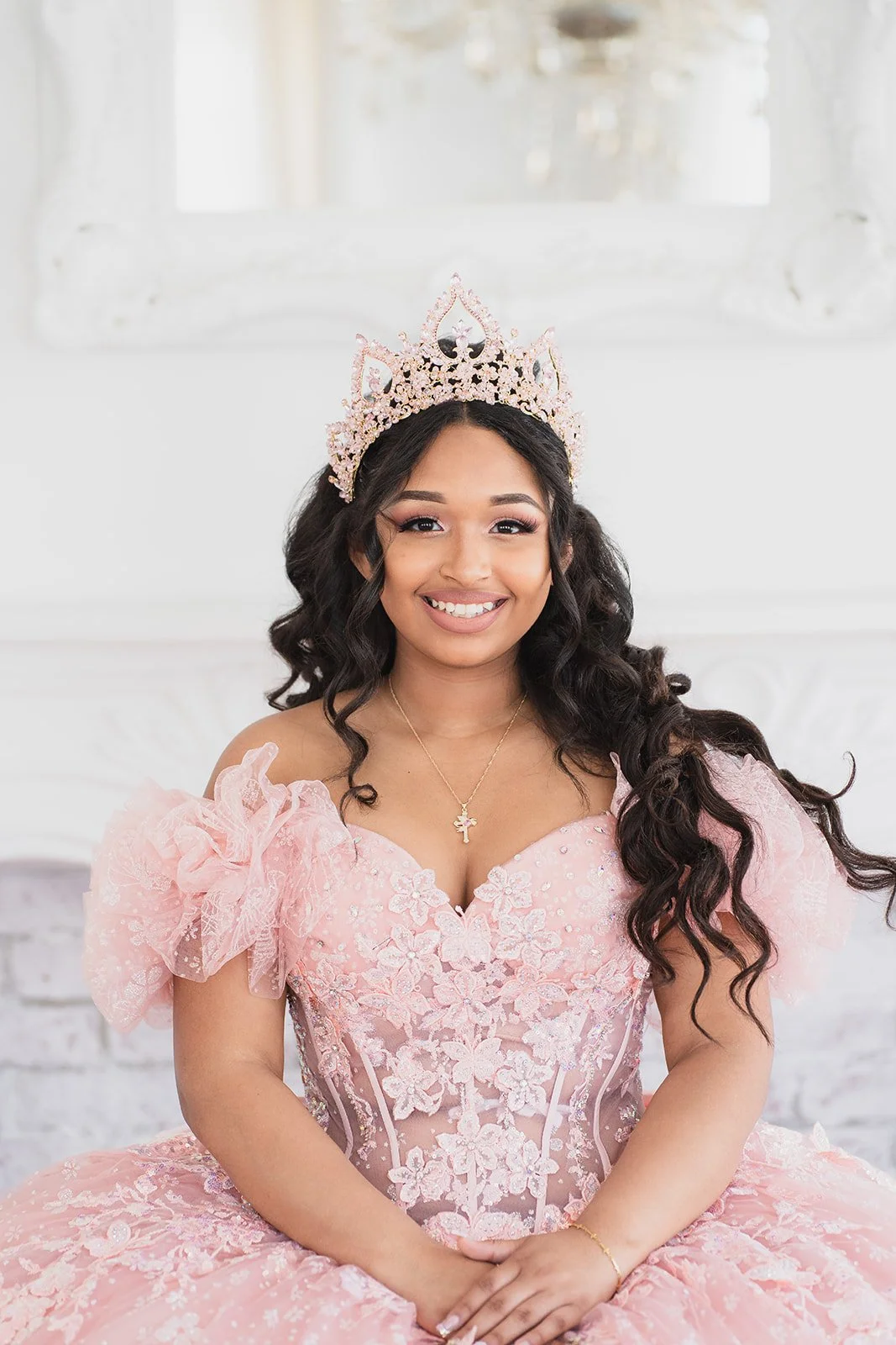 A young woman wearing a pink floral lace dress and a tiara, smiling, with loose dark curly hair, in a bright room with a white mirror in the background.