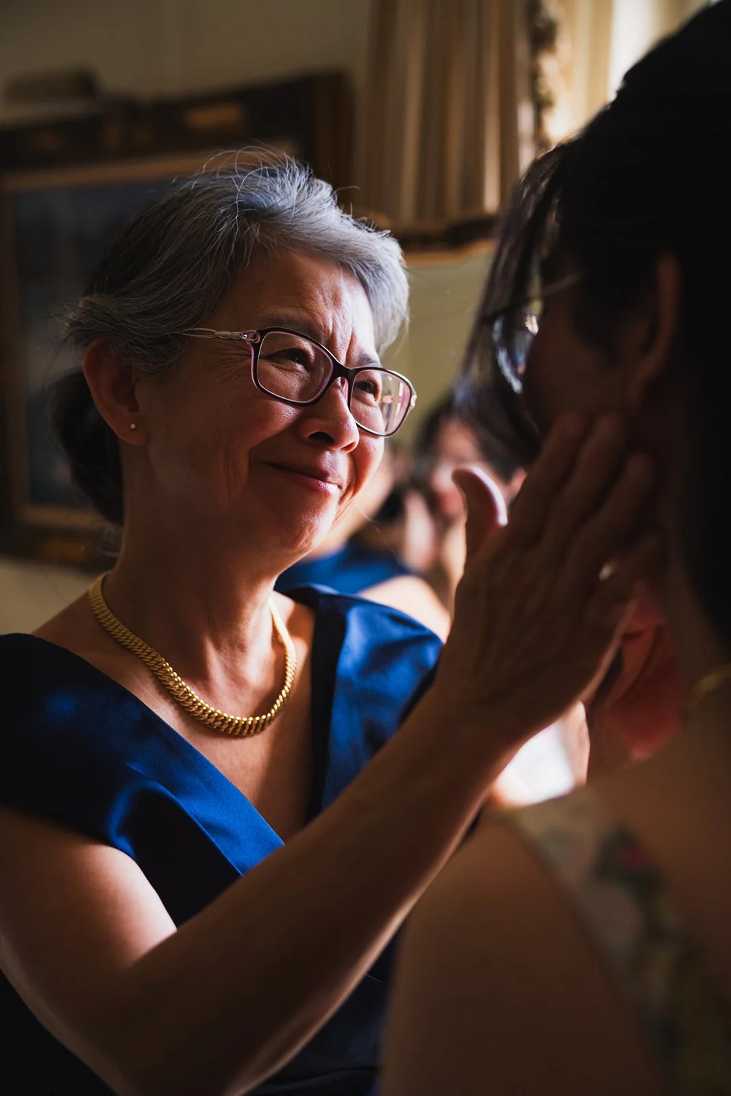 An elderly woman with gray hair, glasses, and a gold necklace smiling at a young woman, engaging in a warm, emotional moment indoors.