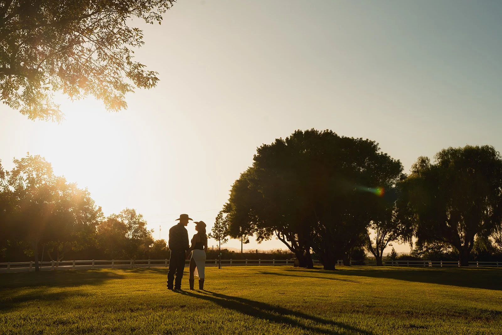 A man and woman standing close together outdoors on a grassy field during sunset, holding hands and facing each other, with large trees and a white fence in the background.