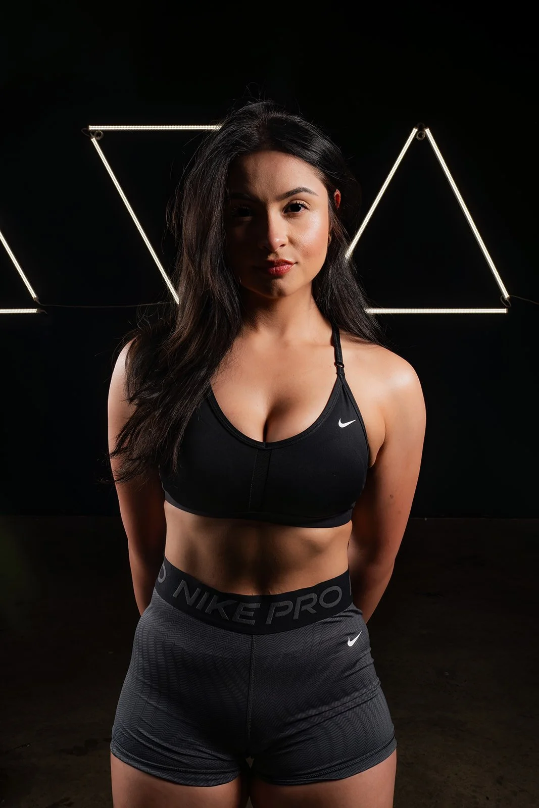 A fit woman in black Nike sports bra and gray Nike Pro shorts standing against a black background with illuminated geometric shapes behind her.