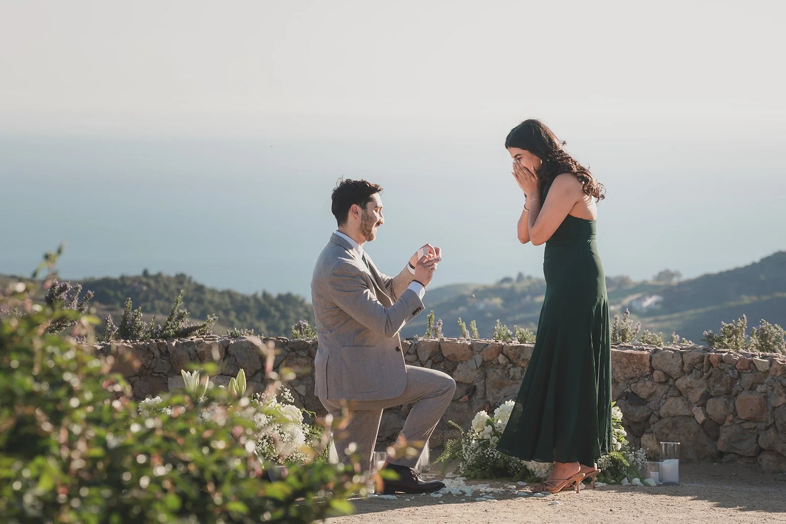 A man down on one knee proposing, with a woman covering her face with both hands in excitement and shock, on a mountain top overlooking Malibu