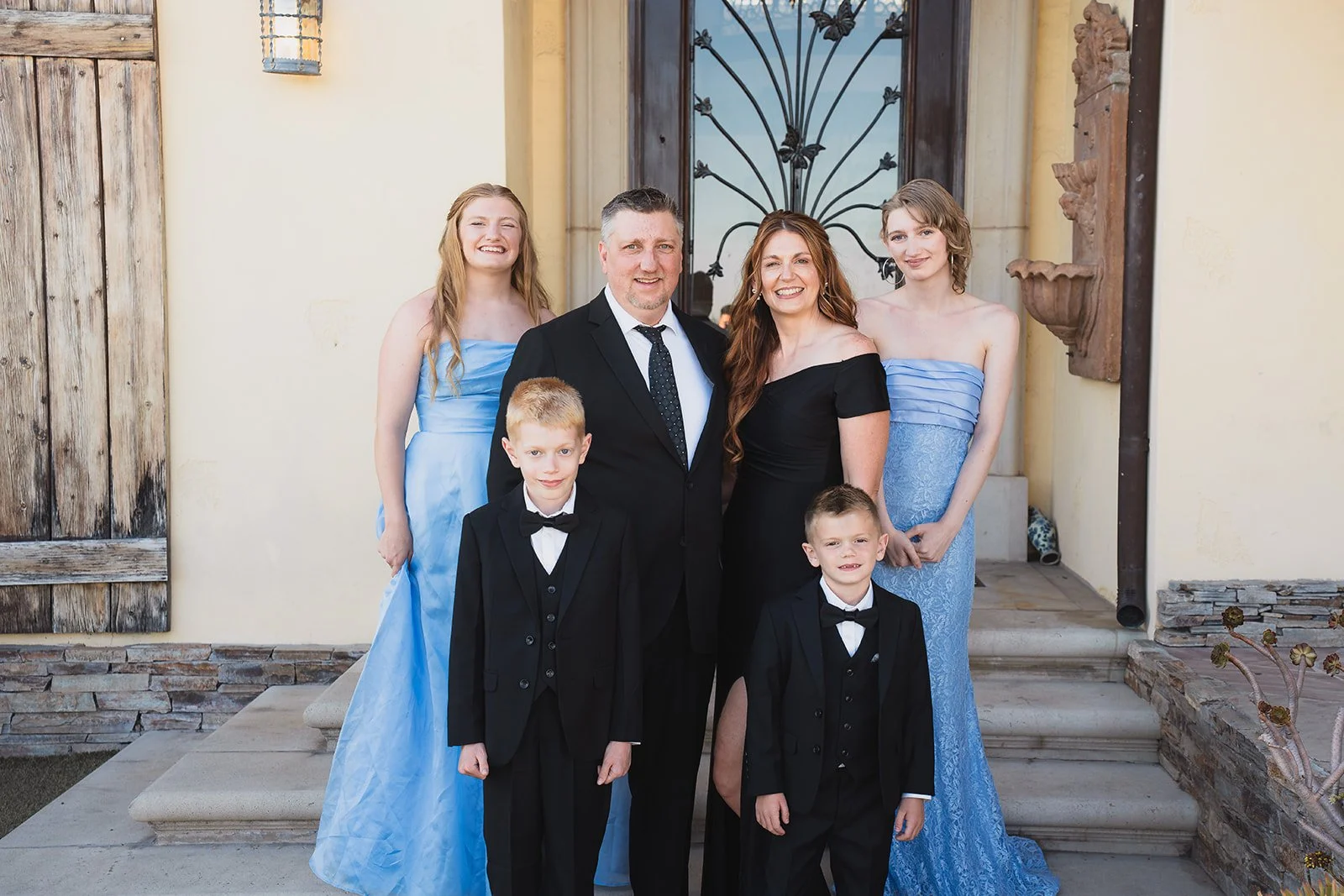 A family of six dressed in formal attire posing on the steps outside a house with a wooden door and decorative ironwork.