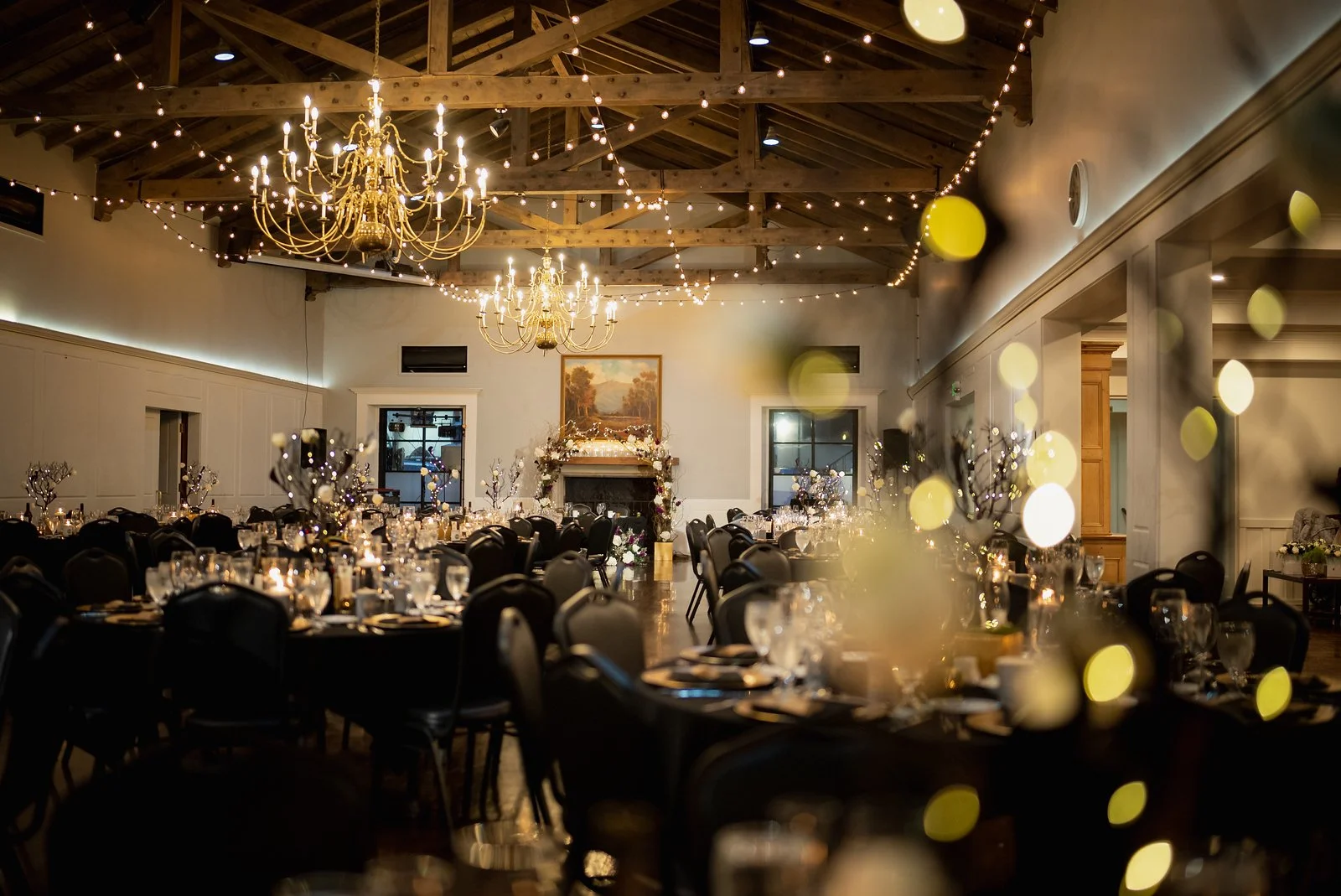 Elegantly decorated banquet hall with tables set for a formal event, chandeliers hanging from a wooden vaulted ceiling, and string lights creating a warm ambiance.