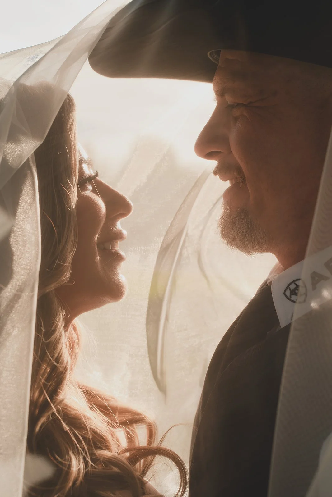 A bride and groom face to face behind a veil, smiling at each other with backlit soft sunlight.