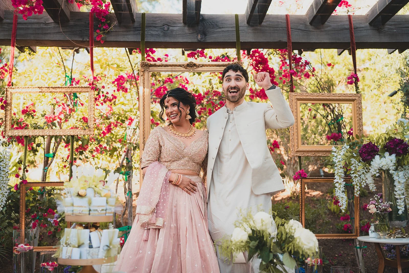 A couple at their wedding celebration, standing in front of a decorated floral backdrop with empty golden frames, with bouquets and wedding cake on display.