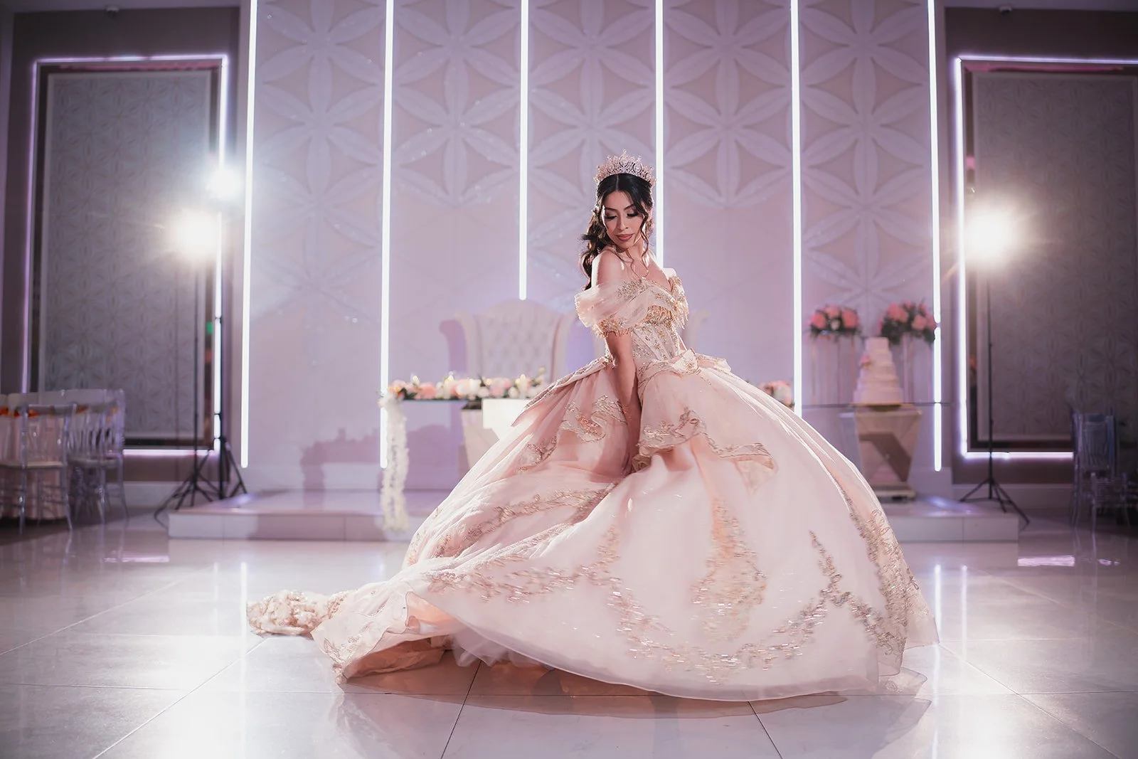 A woman in a pink, elaborate ball gown with gold embroidery, wearing a tiara, posing in a decorated event hall with flowers and a stage in the background.