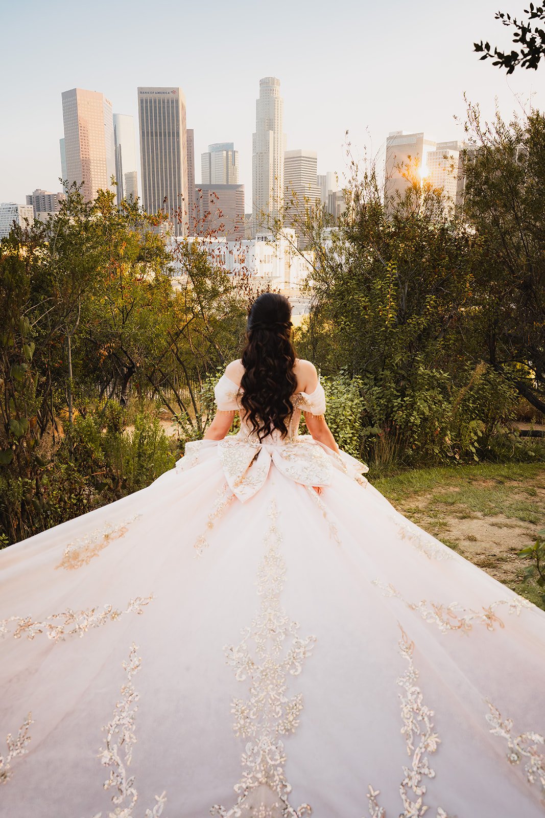 A woman in an elegant wedding gown with a full, embroidered train stands outdoors, facing a city skyline at sunset.