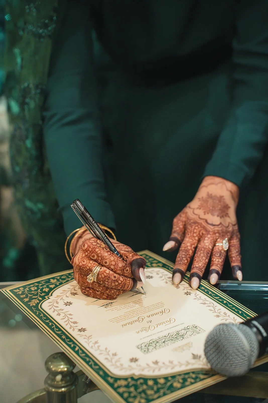 A person with intricate henna designs on their hands is signing a certificate or document with a black pen. The person has multiple rings and a gold bracelet, and there is a microphone on the table nearby.