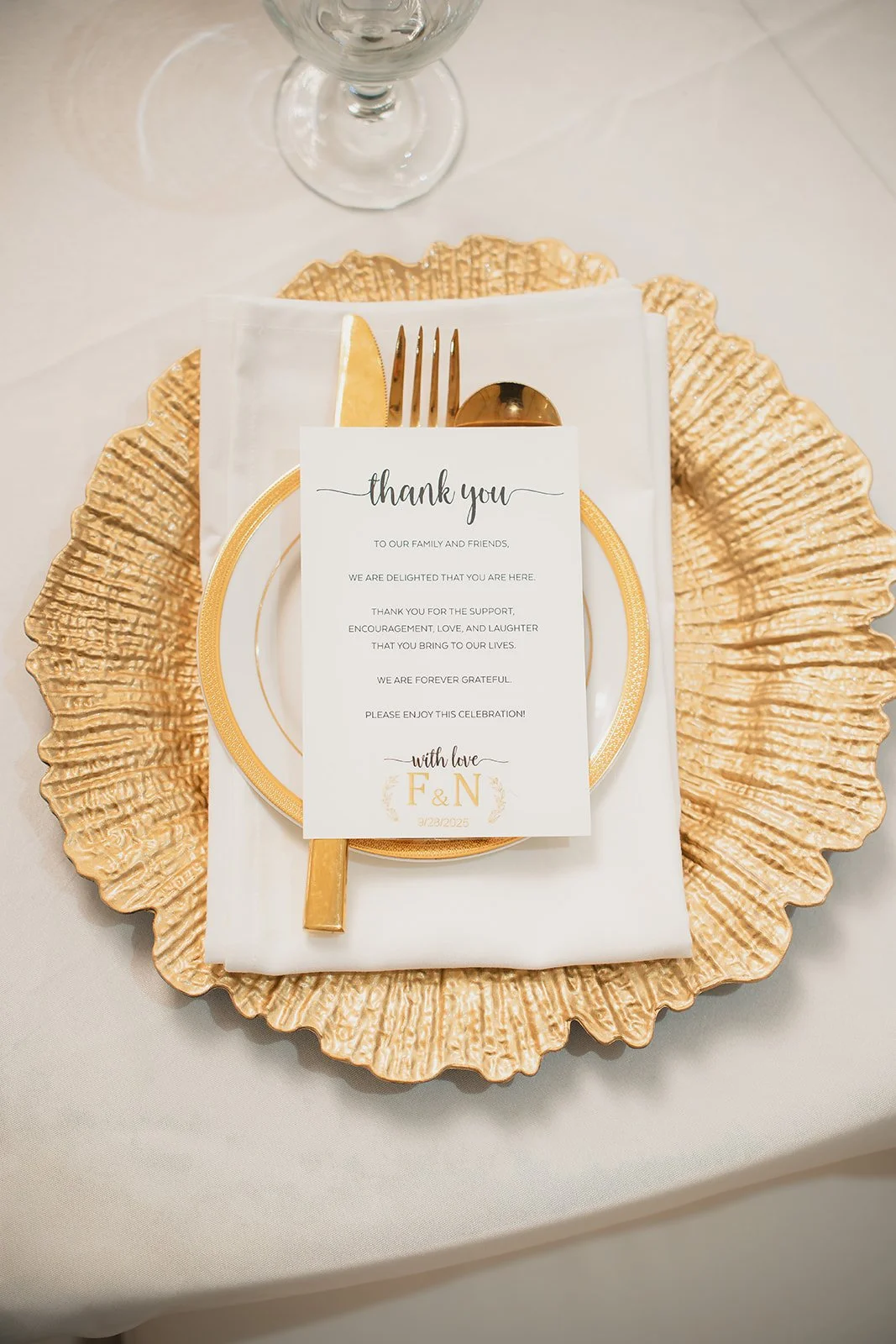 Elegant table setting with a gold textured charger plate, white napkin, gold cutlery, and a thank you note on a white card with the initials 'F & N' for a celebration or wedding.