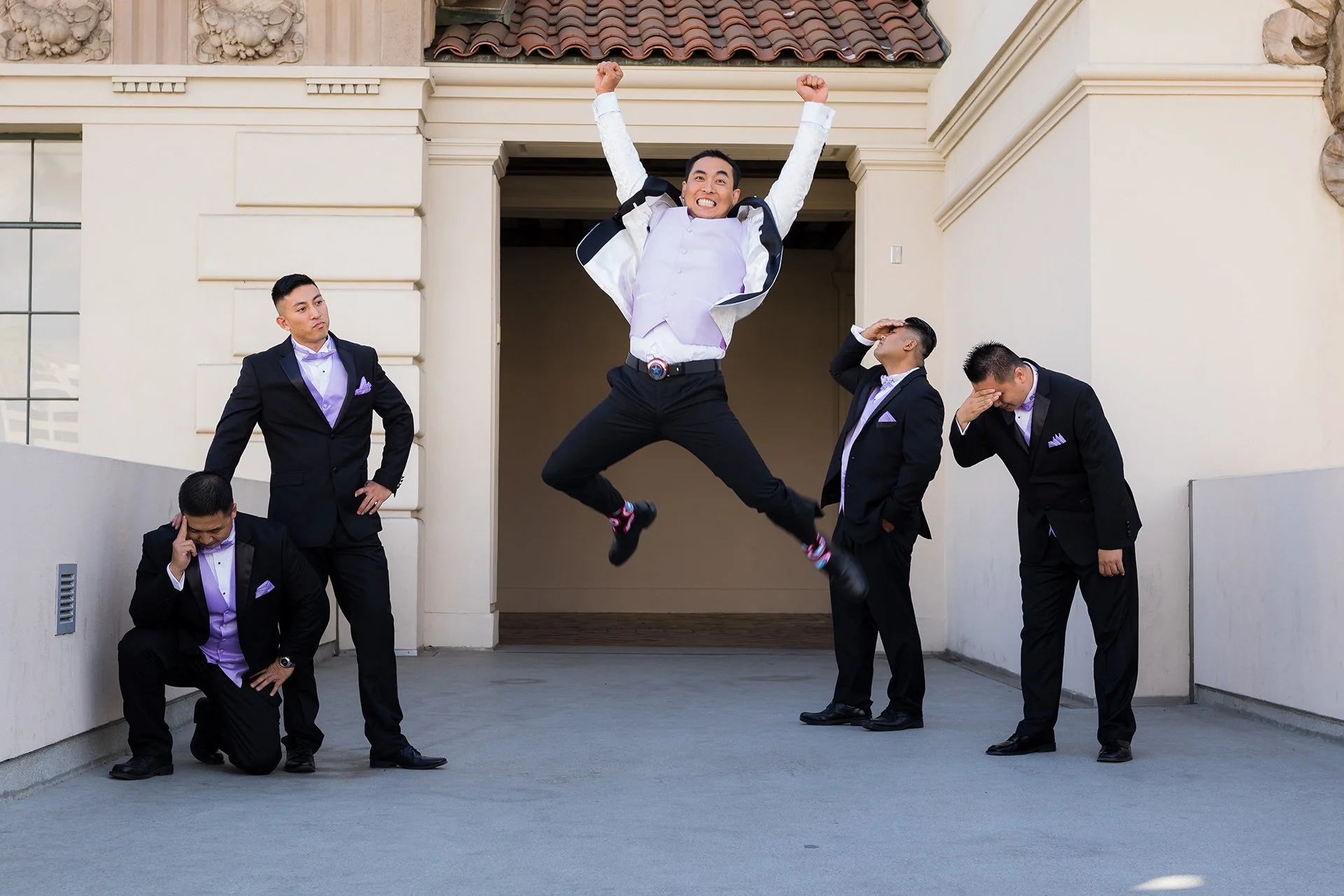 A man in a white shirt and black pants jumps in the air with arms raised, surrounded by five other men in suits, one with a hand on his forehead and another looking down, outside a building with beige walls and a tiled roof.