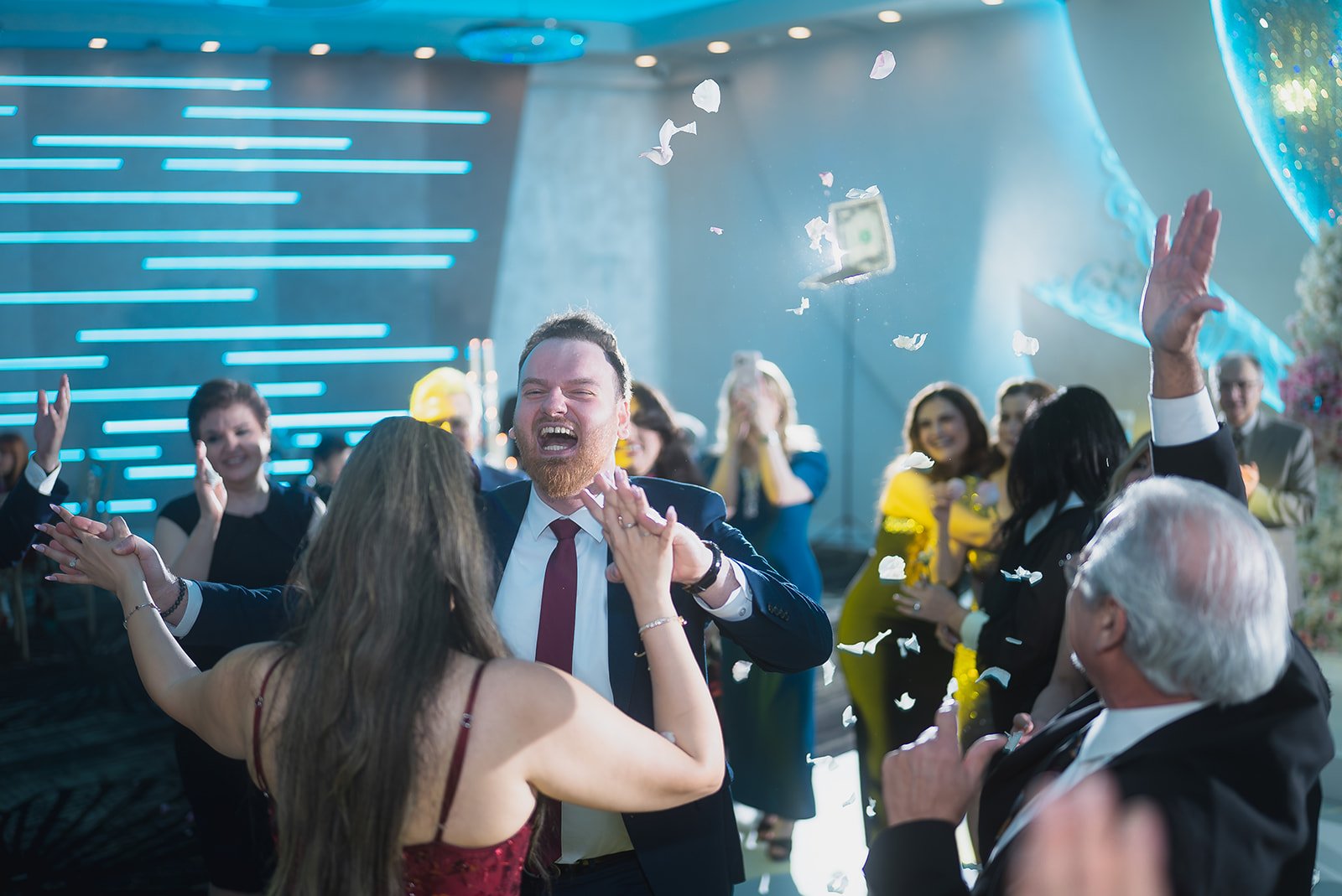 A group of people at a celebration, dancing and throwing confetti in a brightly lit indoor venue.