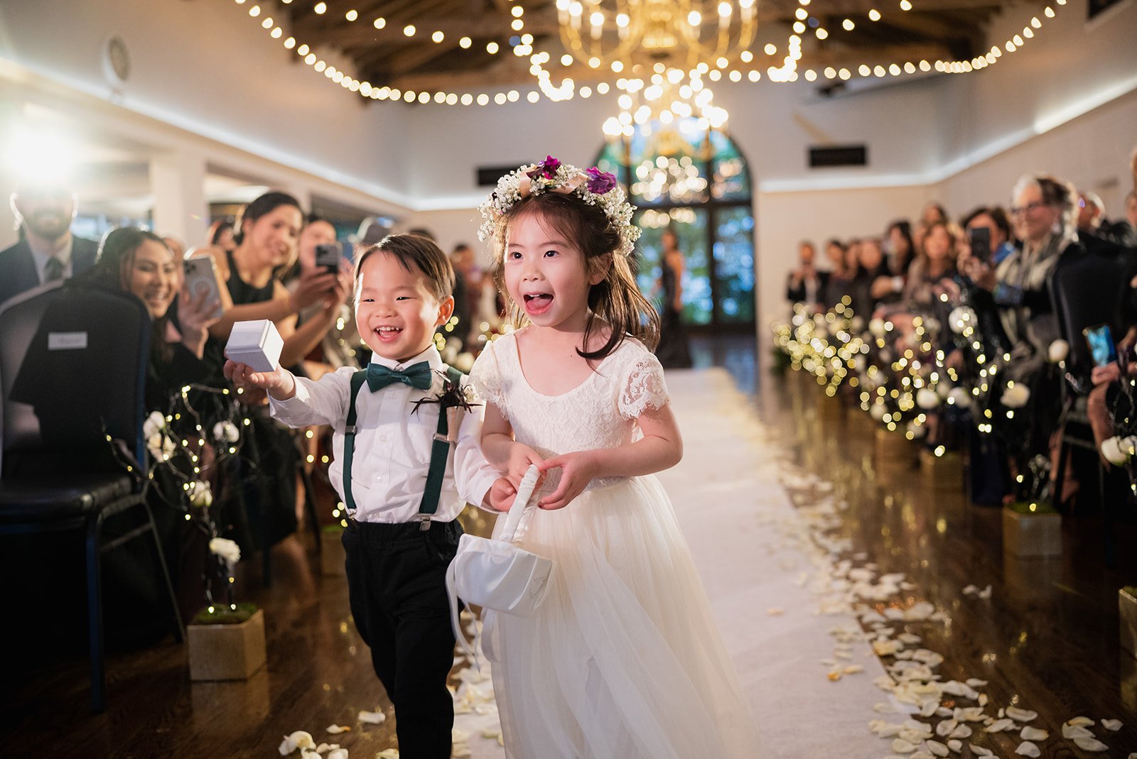 Two young children, a boy and a girl, are walking down an aisle at a wedding, smiling and joyful. The girl is wearing a white dress and a floral crown, and the boy is dressed in a white shirt with suspenders and a bow tie, holding a small box. The ve