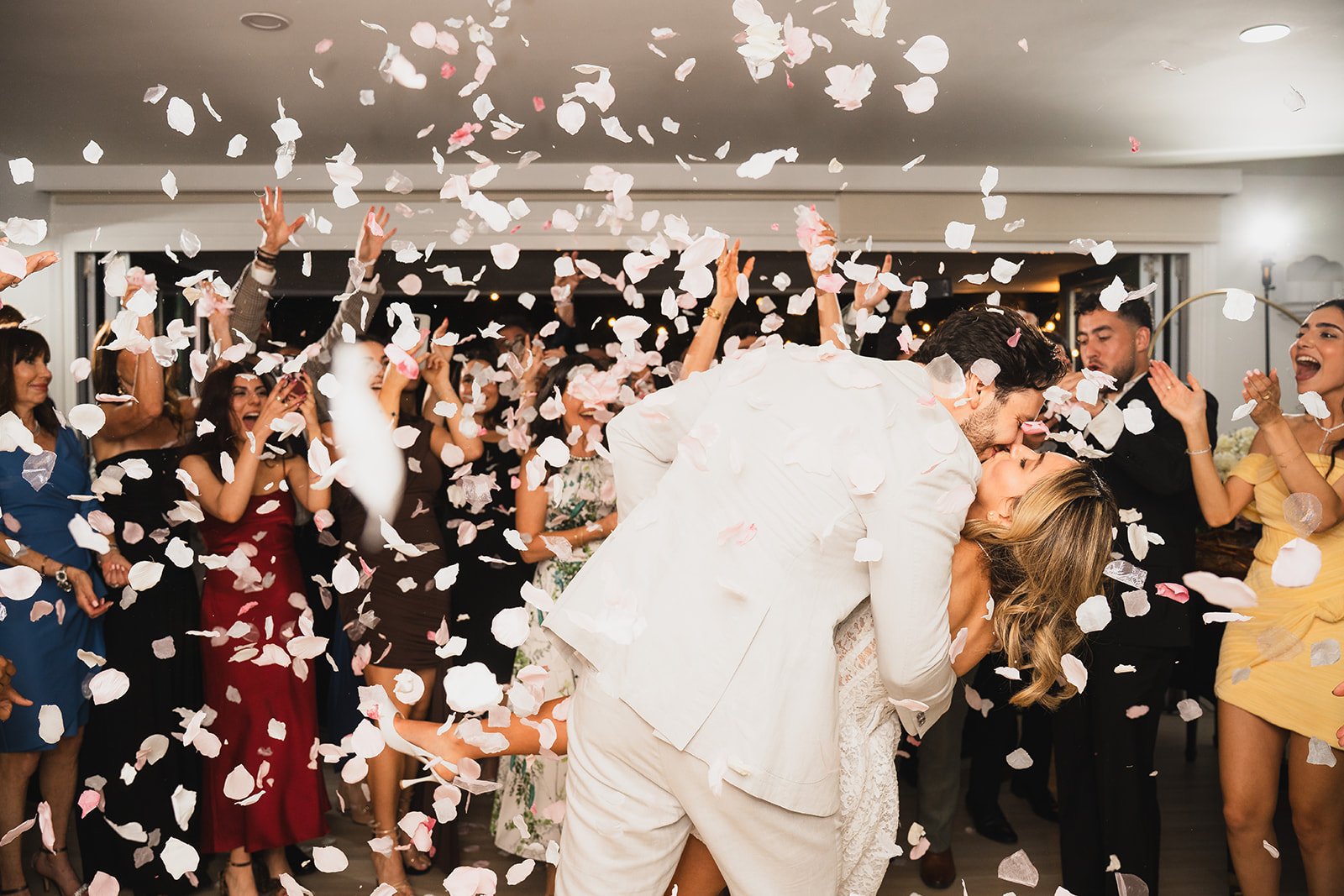 A couple sharing a kiss at a wedding reception, surrounded by guests throwing confetti.