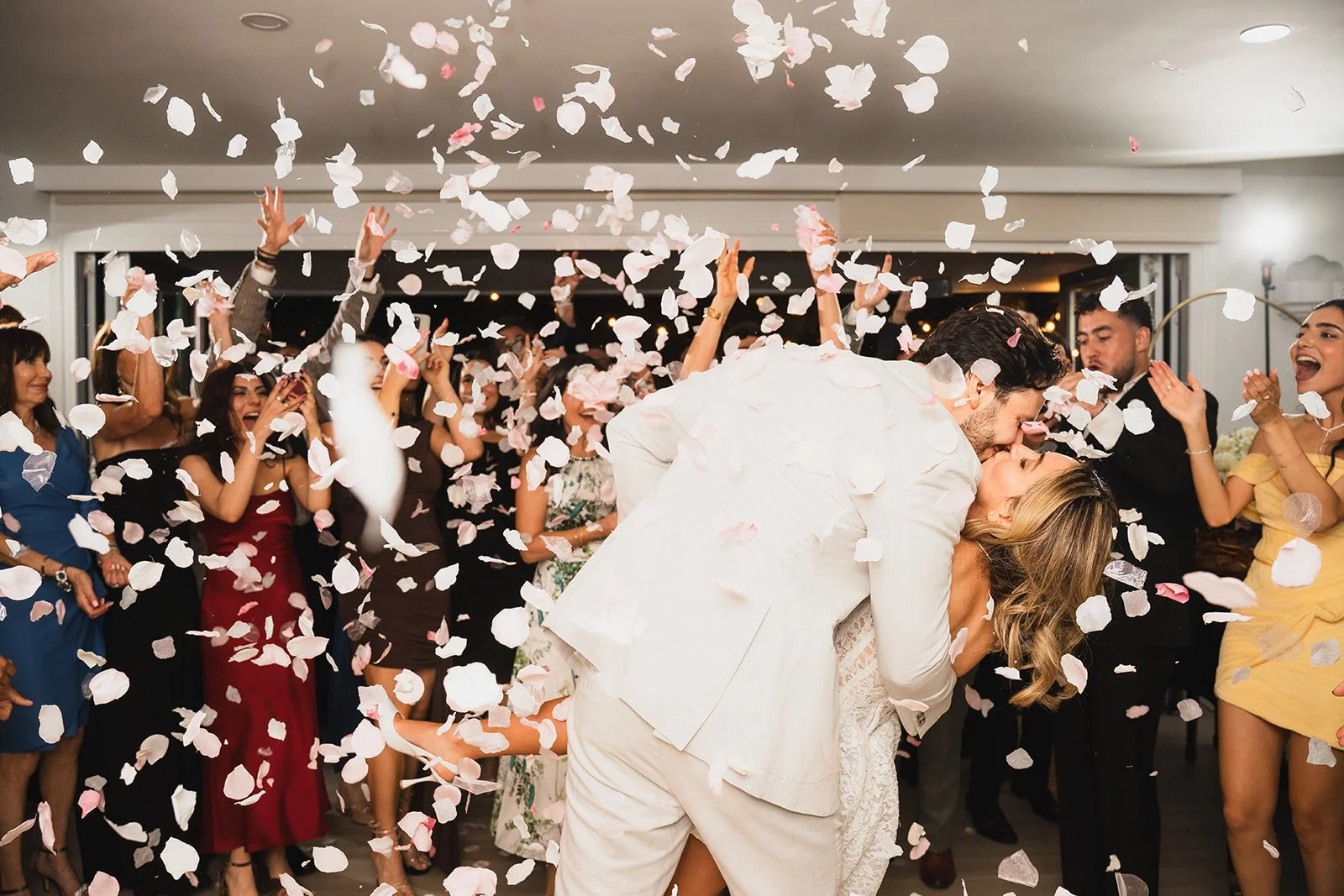 A couple sharing a kiss at a wedding reception, surrounded by guests throwing confetti.