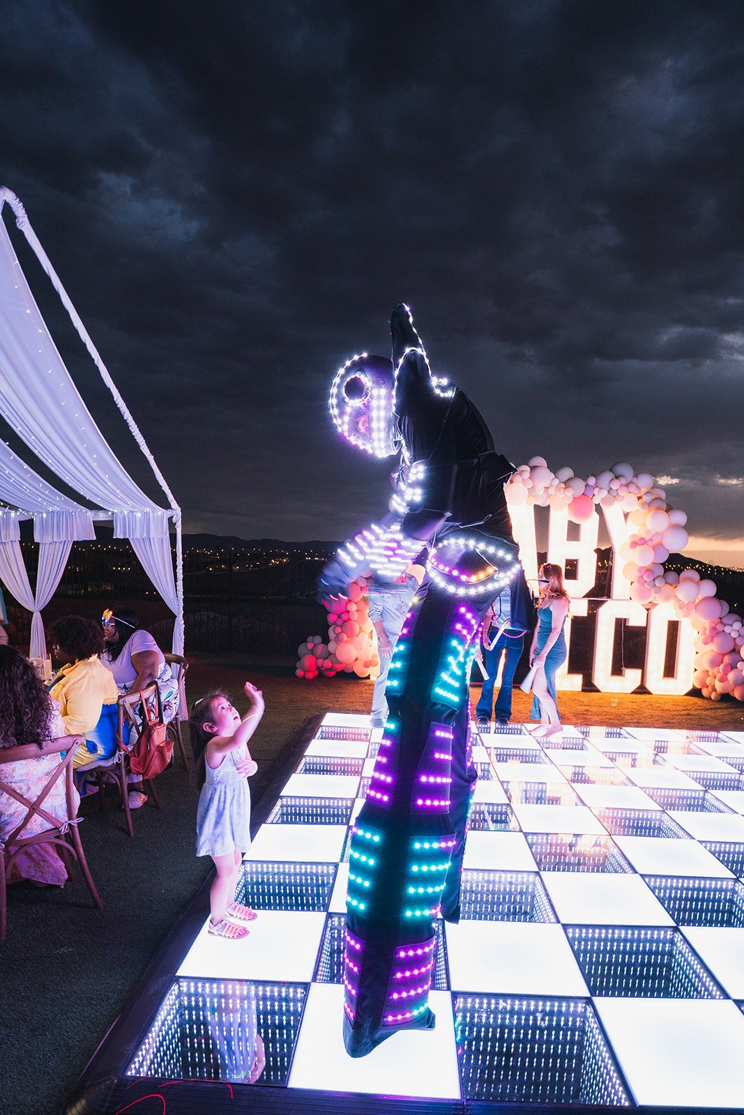 A nightclub scene with a person in a black suit with LED lights performing on a lit-up dance floor. A young girl is reaching towards the performer, while other guests sit at nearby tables. In the background, balloon decorations spell 'HI ALCO'.