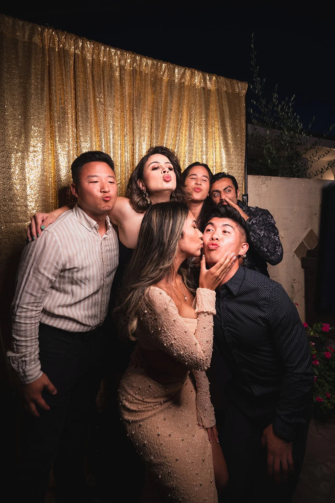 Group of seven young adults at a party making kissing and silly faces, standing close together in front of a sparkly gold backdrop.