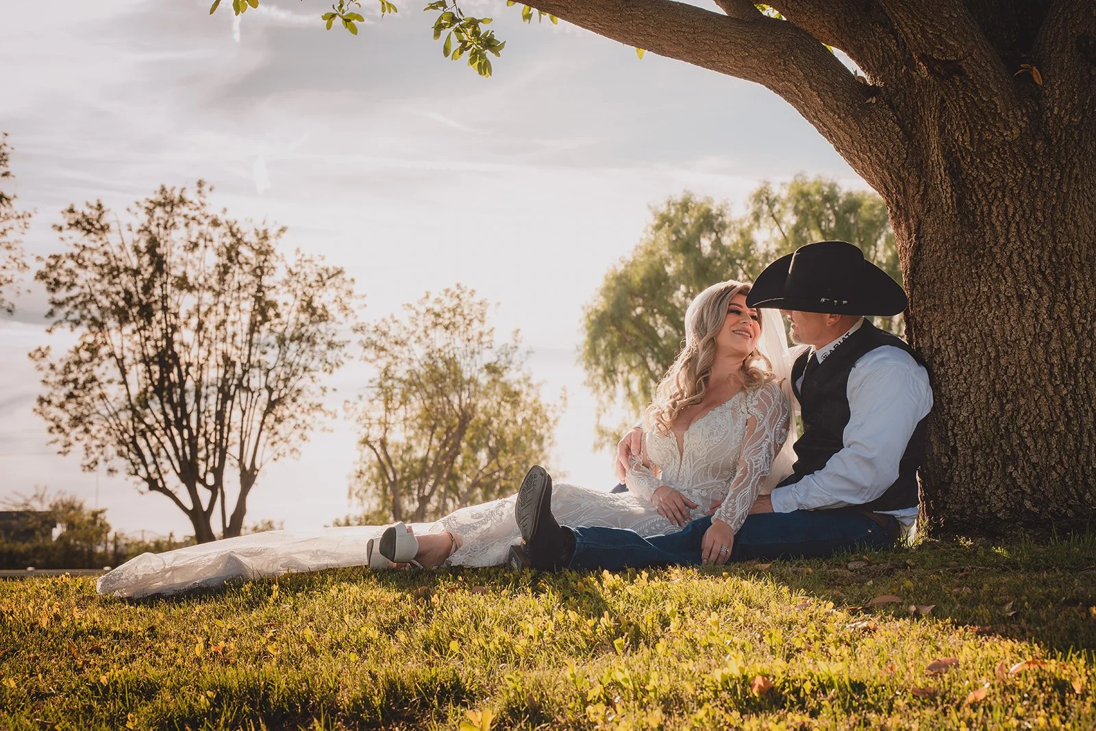 A couple sitting under a large tree on grass, smiling and looking at each other. The woman is in a wedding dress, and the man is in a cowboy hat and western attire. Sunset lighting creates a warm atmosphere.